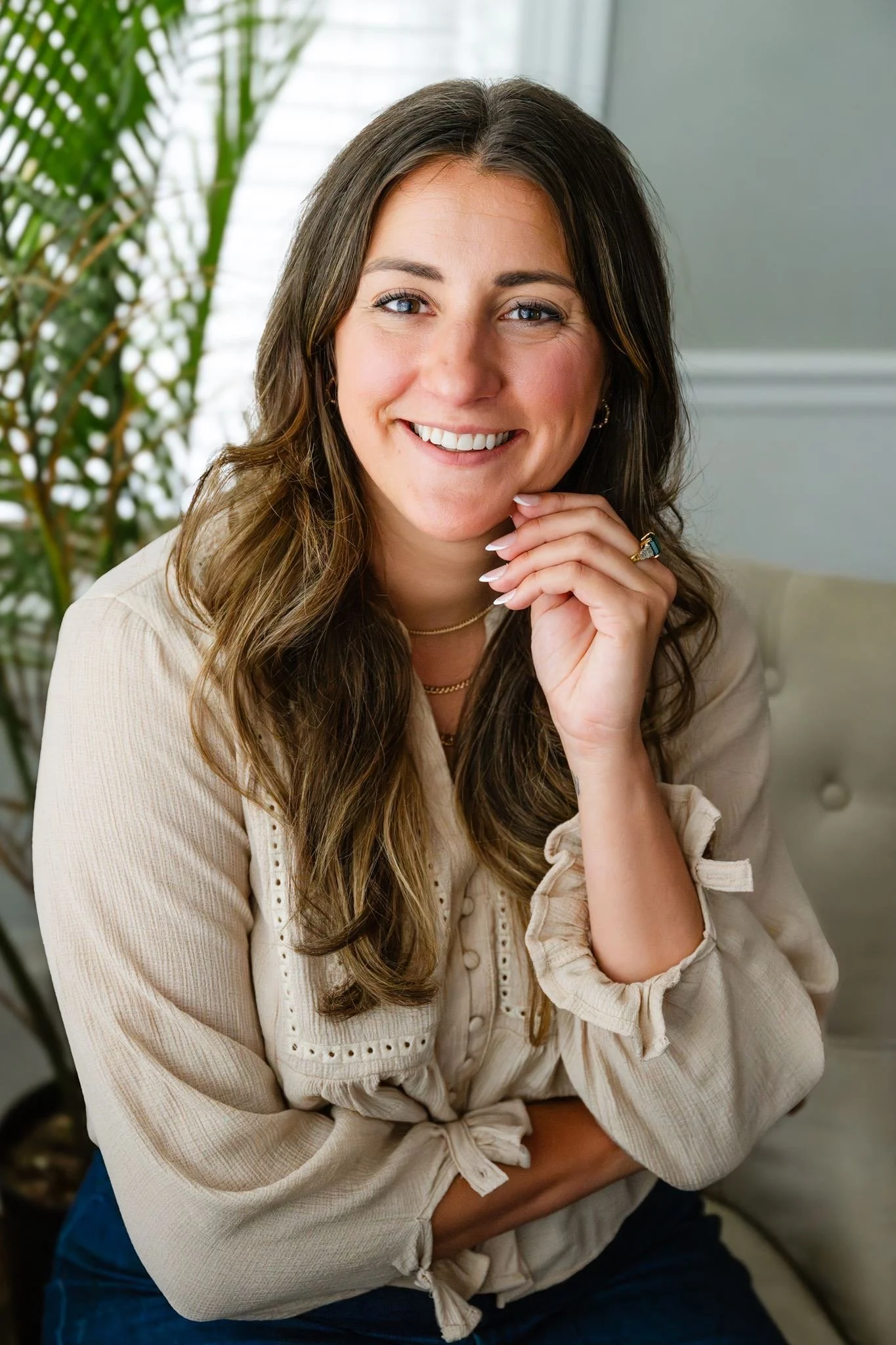 Portrait of a woman with long brown hair, blue eyes, and light skin, smiling with her hand on her chin, dressed in a beige blouse, sitting indoors near a window with green plant leaves in the background.
