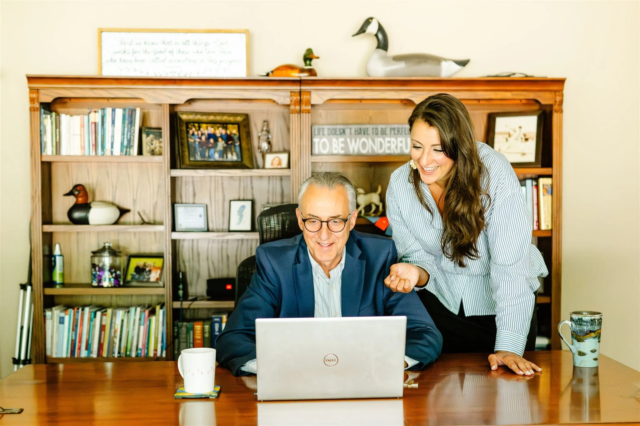 A woman with long brown hair leaning over a man with glasses and gray hair, both sitting at a wooden table and looking at a laptop. The woman is smiling and appears to be pointing at the screen. There are bookshelves with books, framed photos, and decorative items in the background.