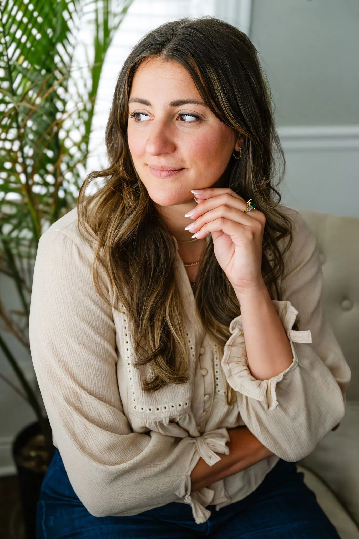 A woman with long brown hair and light makeup, wearing a beige blouse and gold jewelry, sitting with her hand on her chin, looking to the side, in front of a green plant and a light-colored wall.
