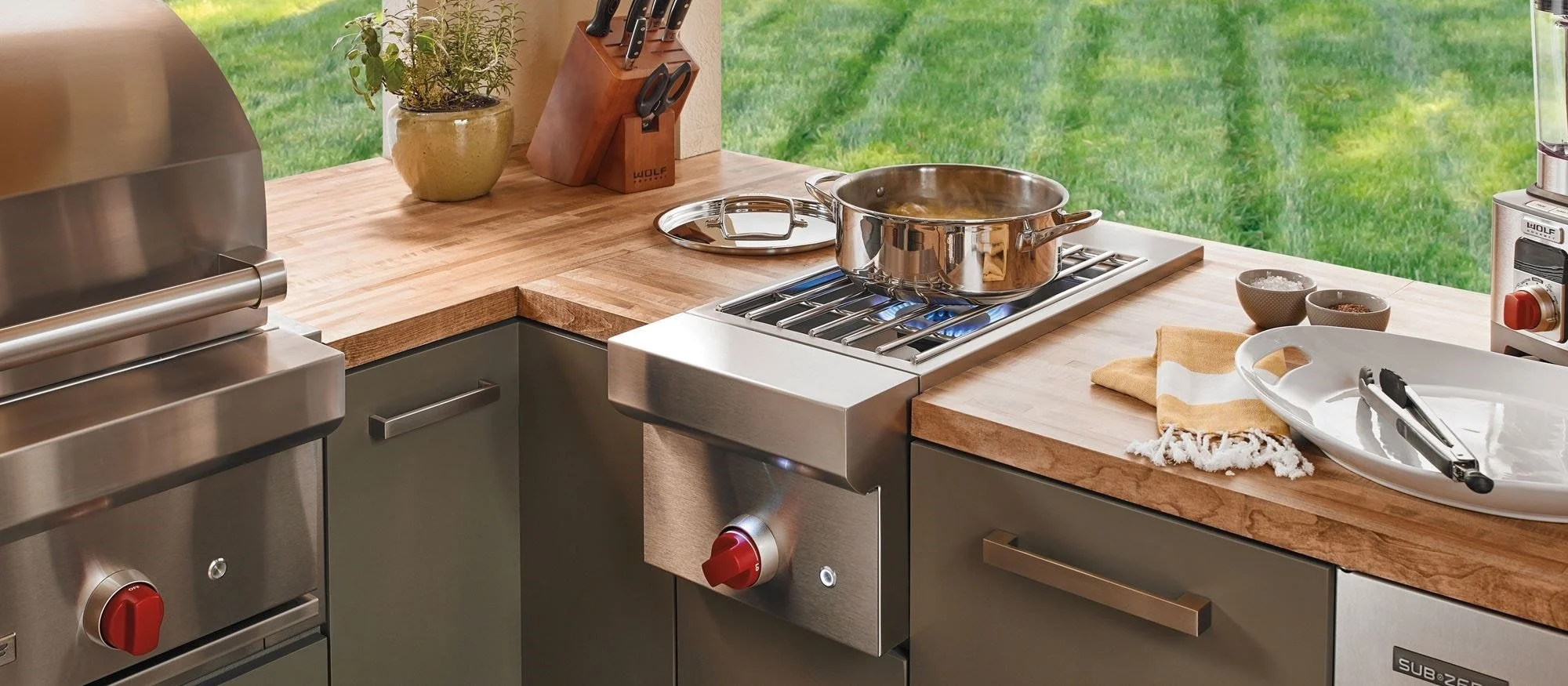 Kitchen countertop with a stove, pot, knives, and dishes, overlooking a grassy backyard.