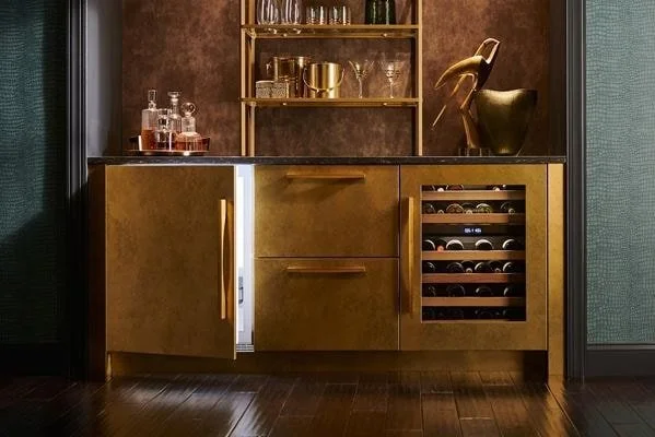 Gold-colored bar cabinet with open wine cooler, shelves with glasses and bottles, and decorative items, against a brown wall and dark wood floor.