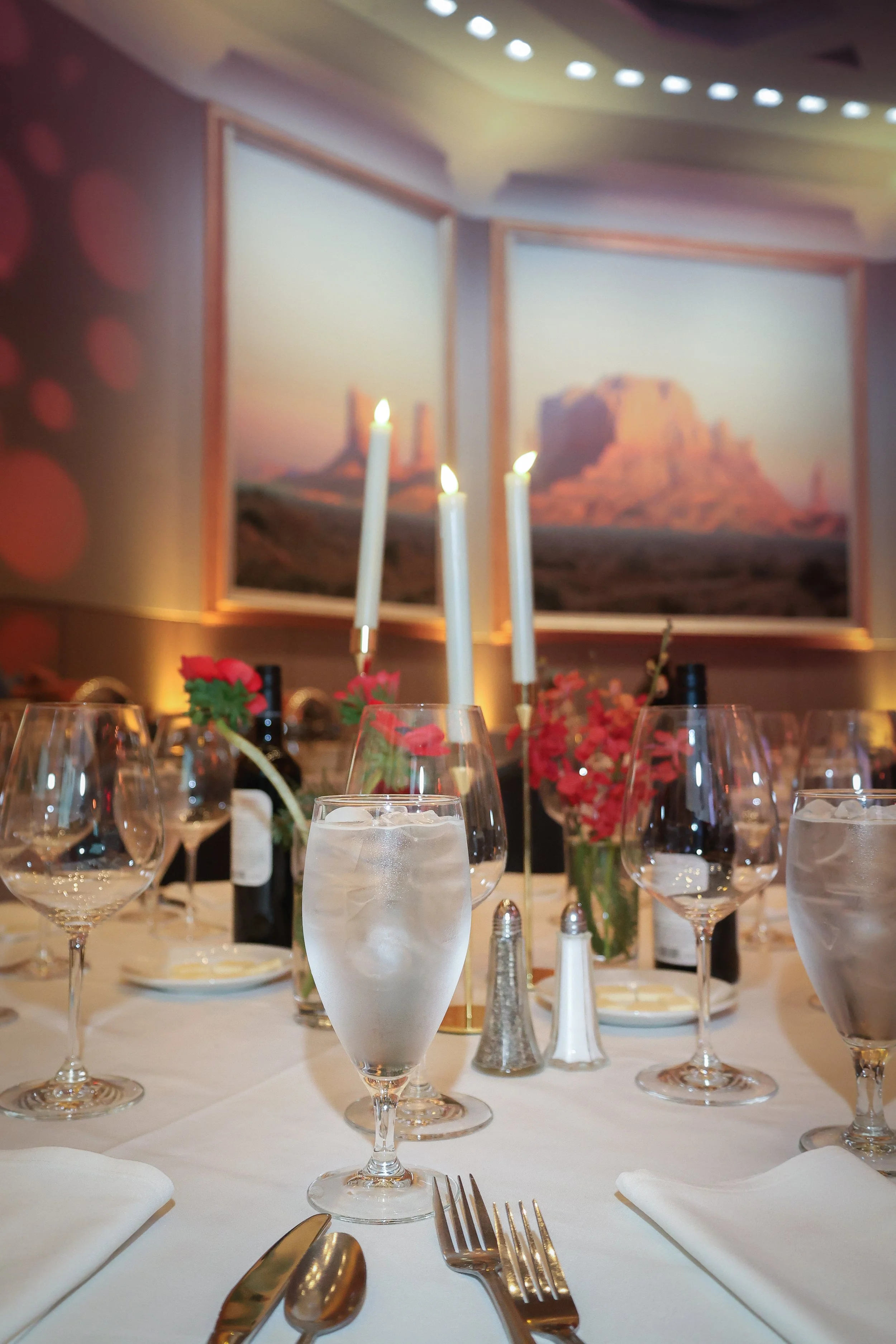Elegant dining table set with water glasses, pink flowers, white napkins, silverware, and white candles, in front of a wall with desert landscape artwork, in a softly lit restaurant.