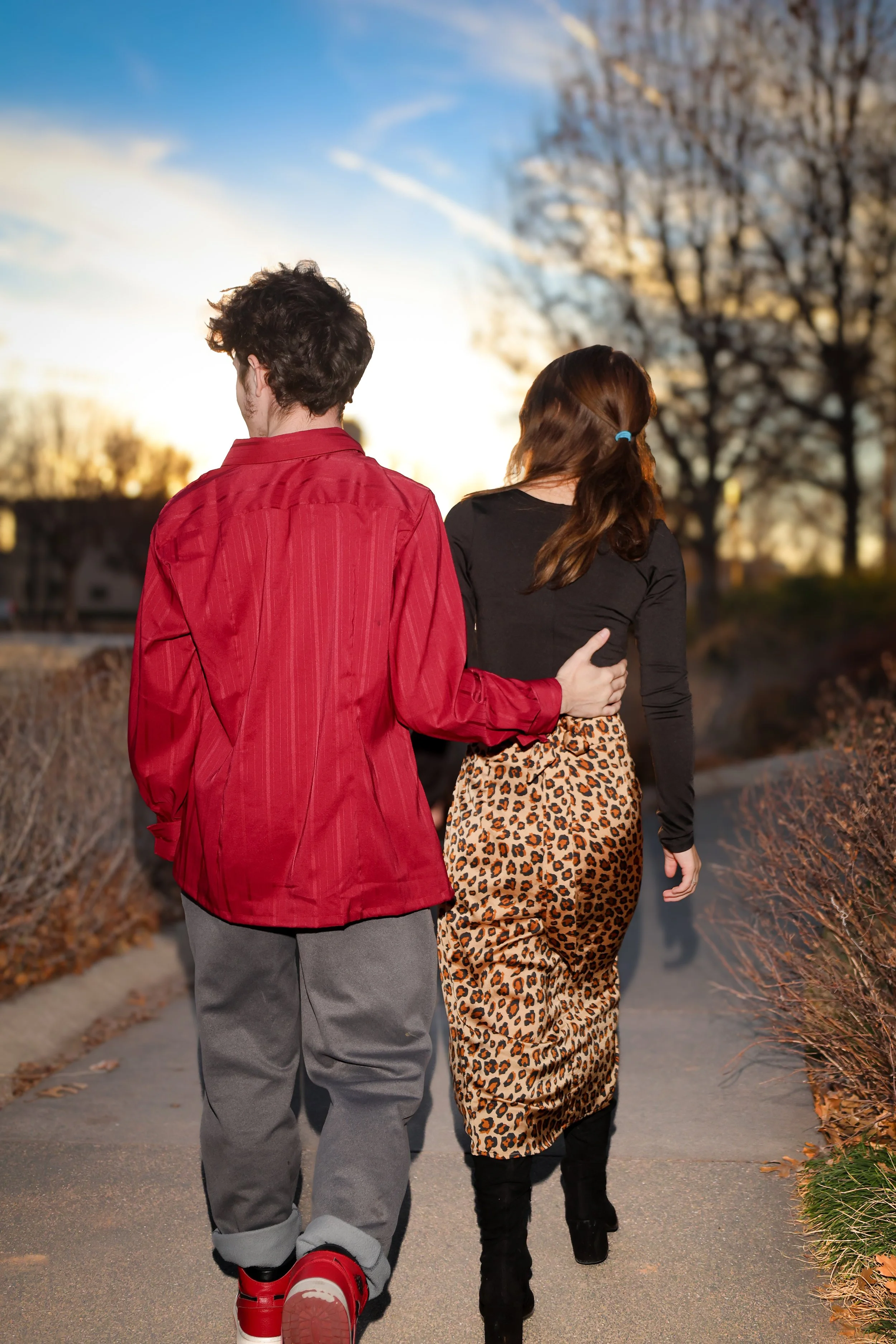 A young couple walking arm in arm on a sidewalk during sunset, with trees and houses in the background.