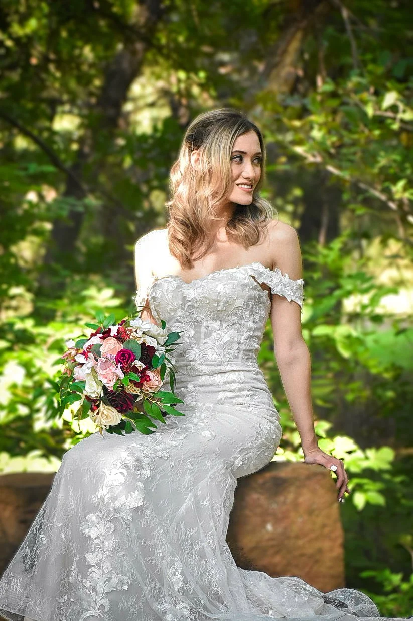 A woman in a white wedding dress sitting on a large rock in a forest, holding a bouquet of pink, white, and red roses with green leaves, looking to her right.
