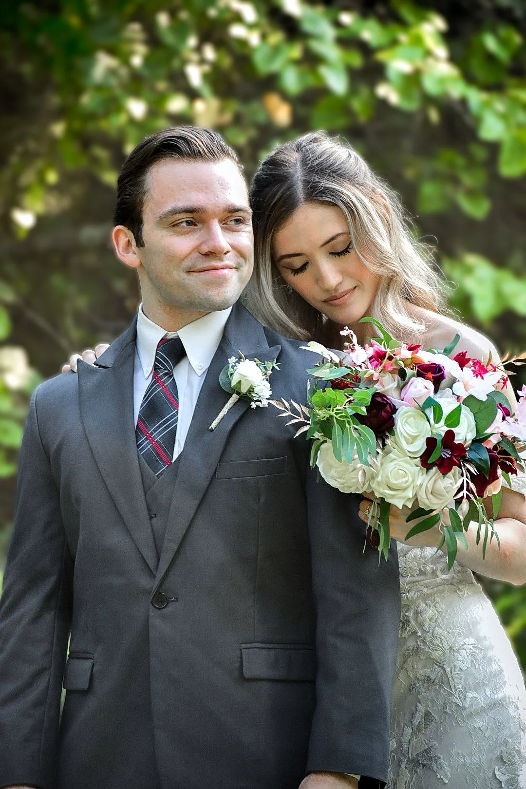 A bride and groom stand outdoors against a background of green trees. The groom, in a dark suit with a boutonnière, looks confident, while the bride, with closed eyes, leans her head on his shoulder and holds a bouquet of white, pink, and burgundy flowers.