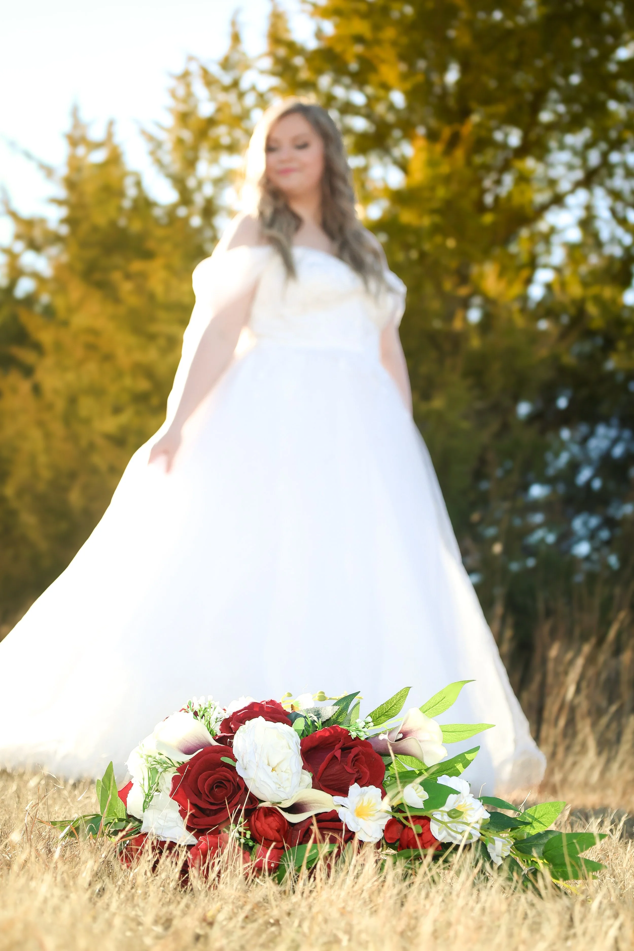 A bride in a white wedding dress standing outdoors with a bouquet of red and white flowers on the grass in front of her, surrounded by trees with autumn-colored leaves.