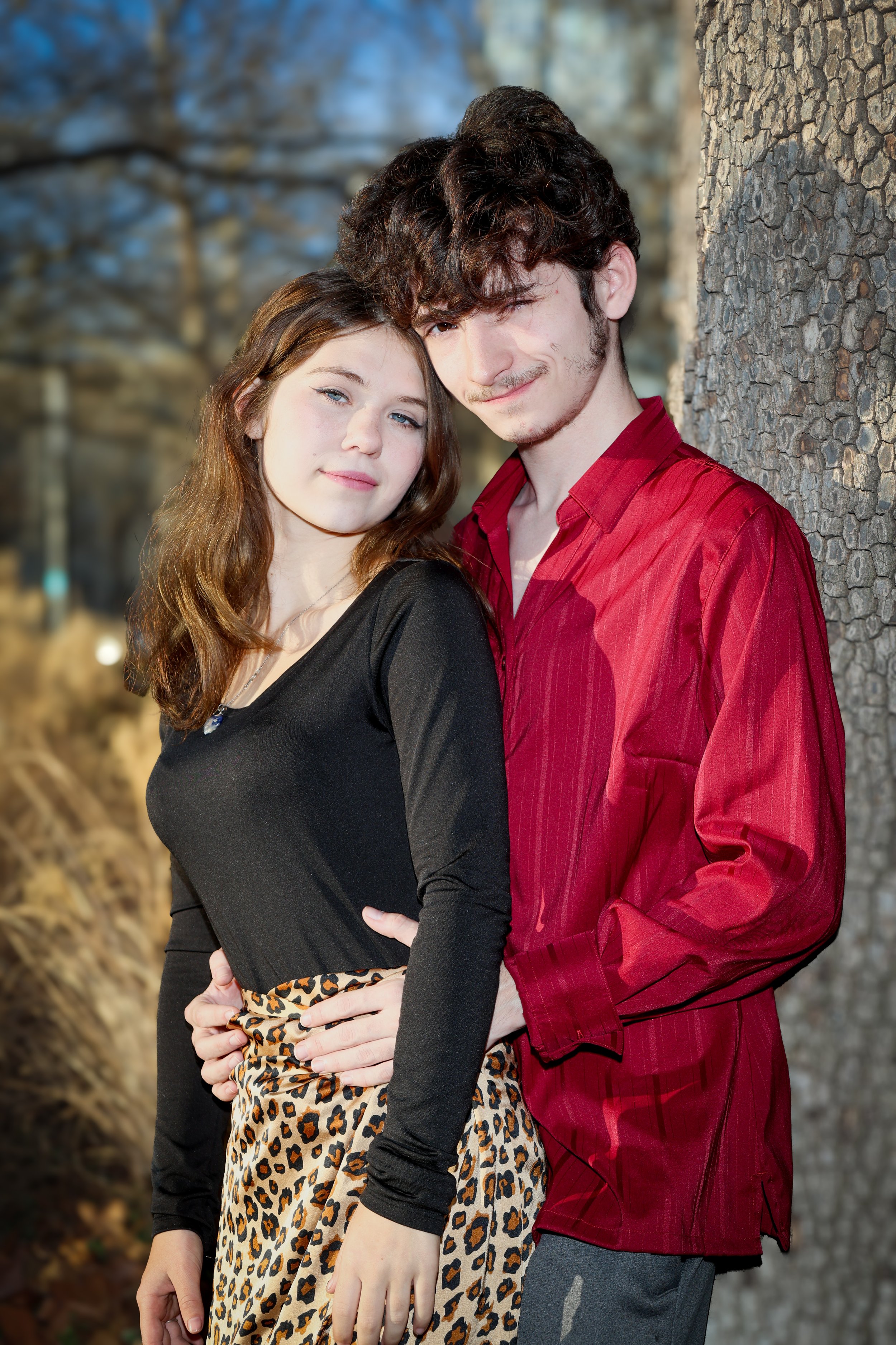 A young couple standing outdoors, leaning against a tree. The woman has long brown hair, fair skin, and is wearing a black top and leopard print skirt. The man has curly dark hair, fair skin, and is wearing a red shirt. They are close together, with 