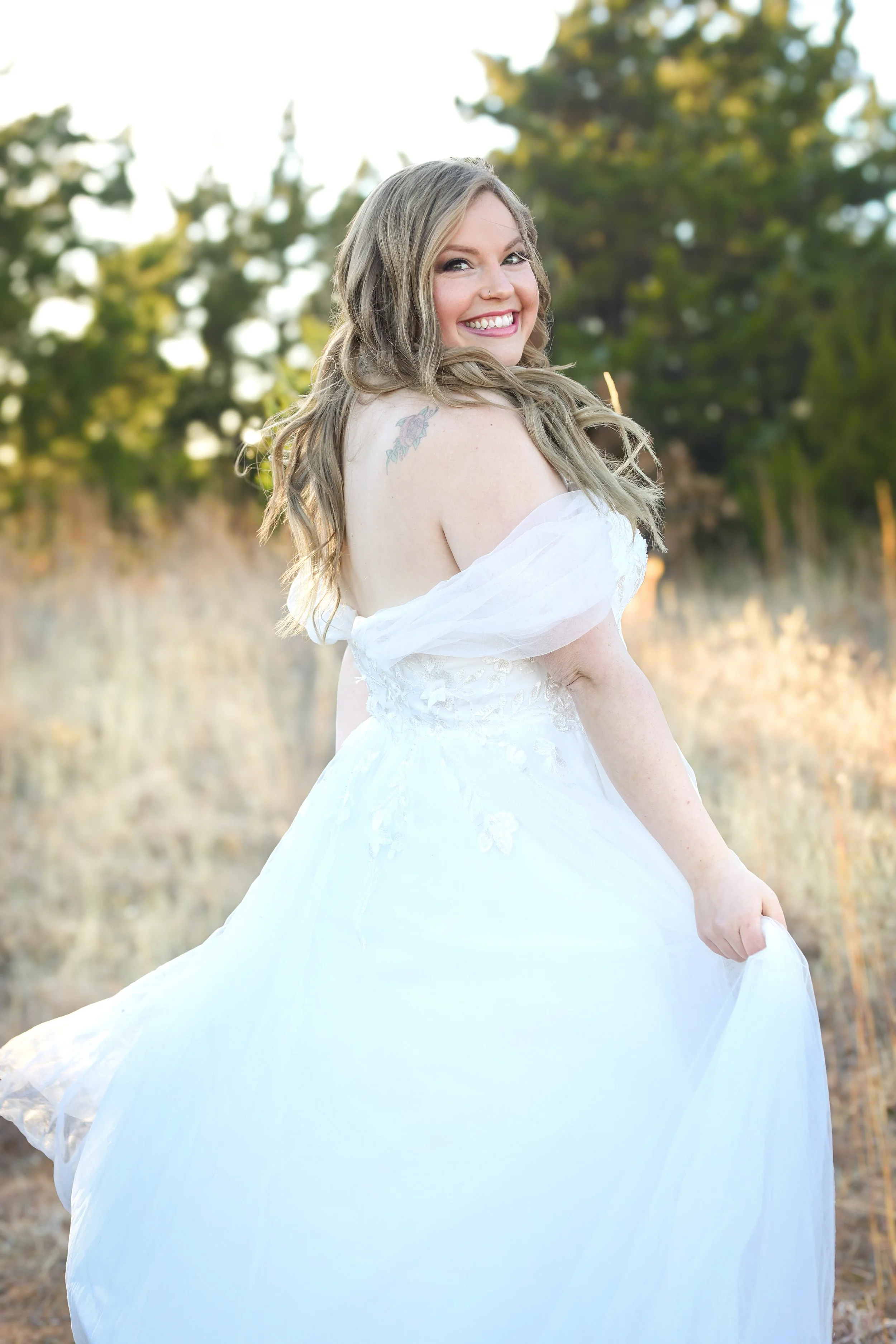 A smiling woman in a white wedding dress outdoors during daytime, holding the skirt of her dress, with trees in the background.