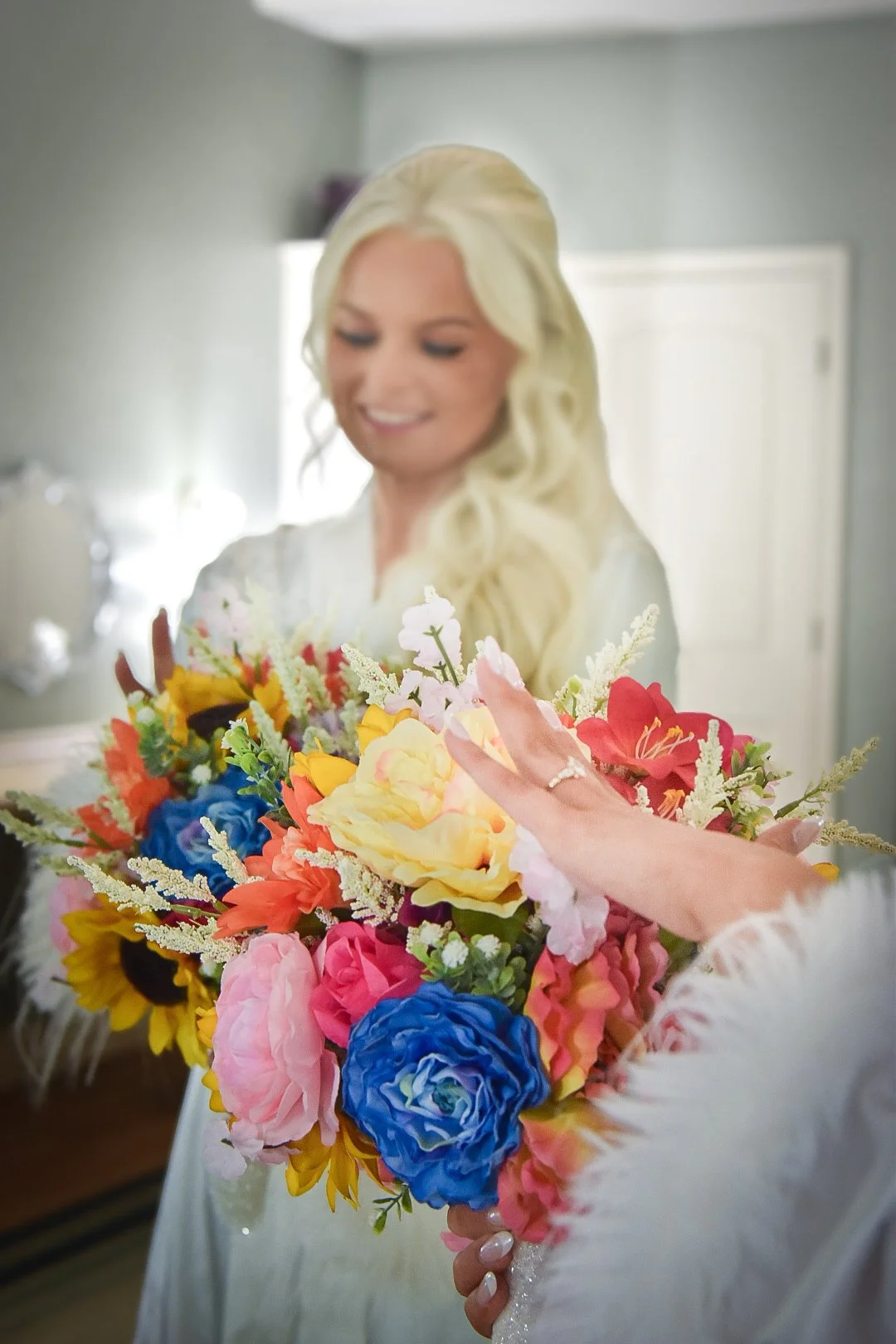 A bride with long blonde hair, wearing a white dress, holding a colorful bouquet of flowers with pink, yellow, orange, blue, and white blossoms, smiling softly.