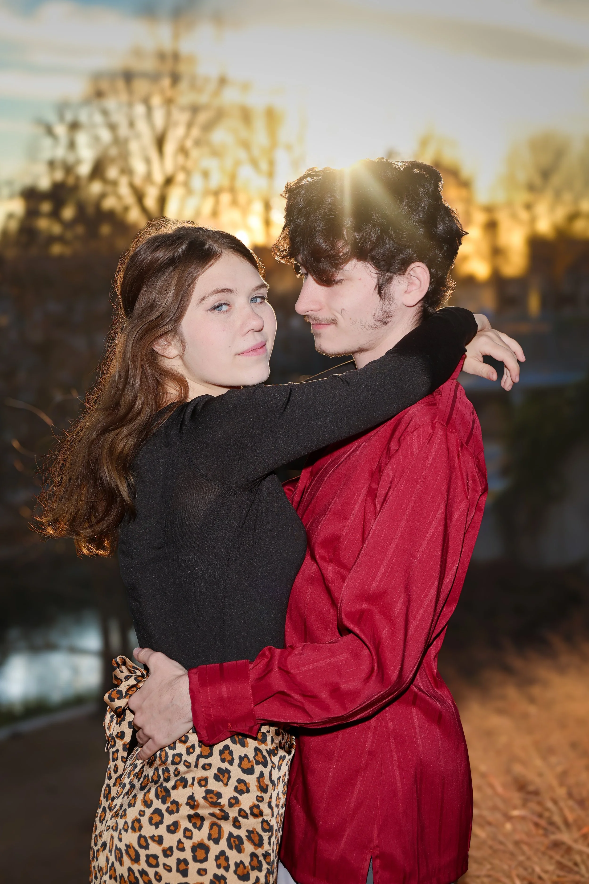 A young couple is hugging outdoors during sunset, with a blurred background of trees and buildings. The woman has long wavy brown hair and is wearing a black long-sleeve top and leopard print pants. The man has curly dark hair, light facial hair, and