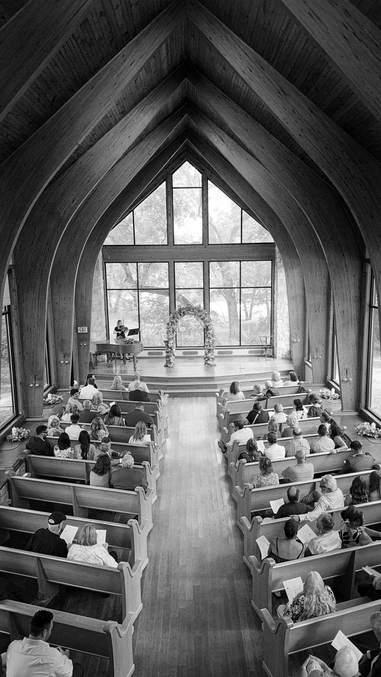 Interior of a wooden chapel with arched ceilings, filled with seated guests attending an event. At the front, a stage with a floral arch and a pianist playing a grand piano.
