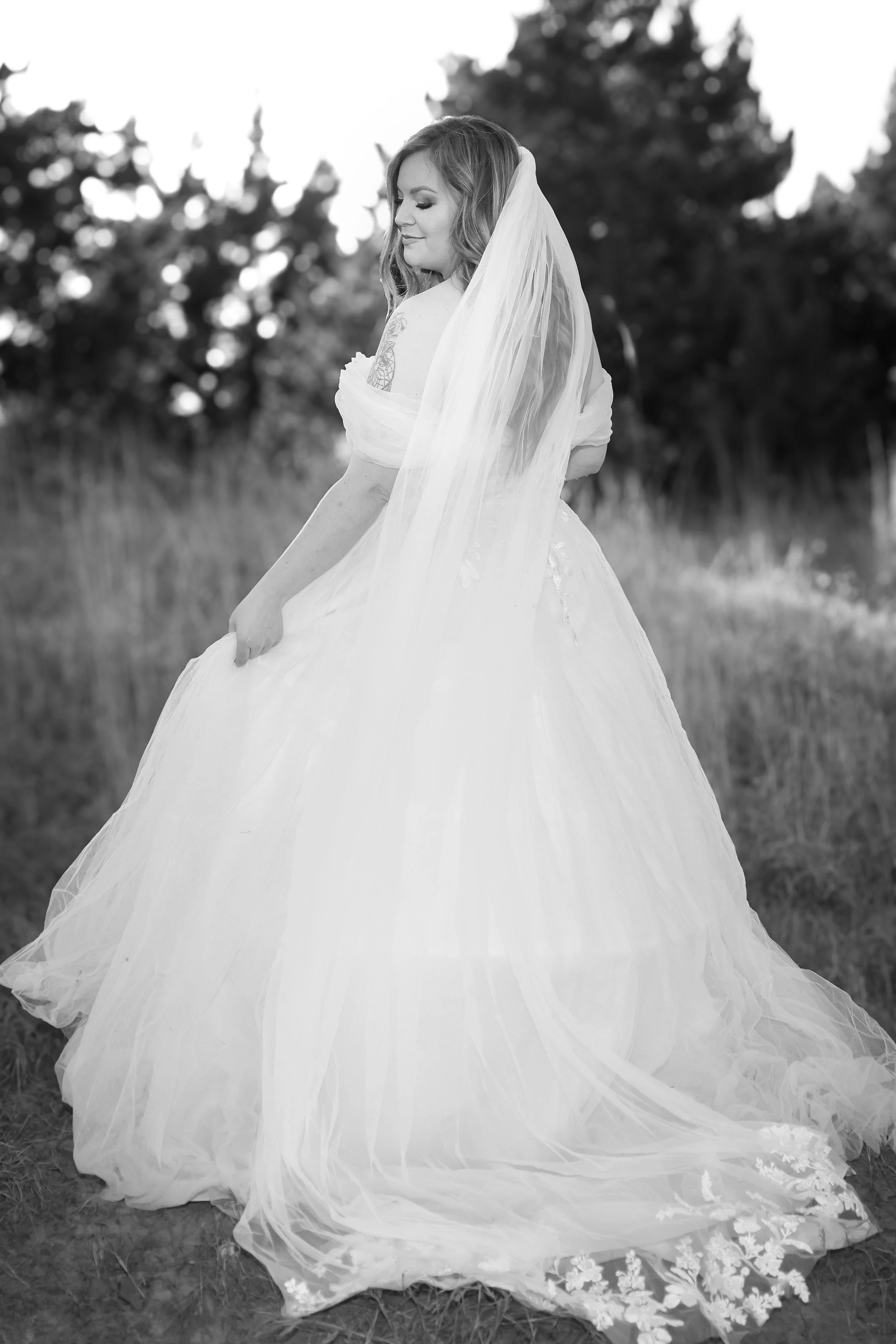 A woman in a wedding dress with a long veil standing outdoors in a grassy area with trees in the background.