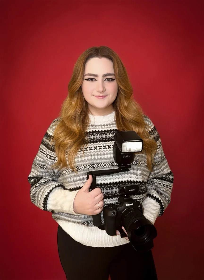 A young woman with long wavy red hair, wearing a black and white patterned sweater, holding a professional camera with a flash attachment, standing against a red background.