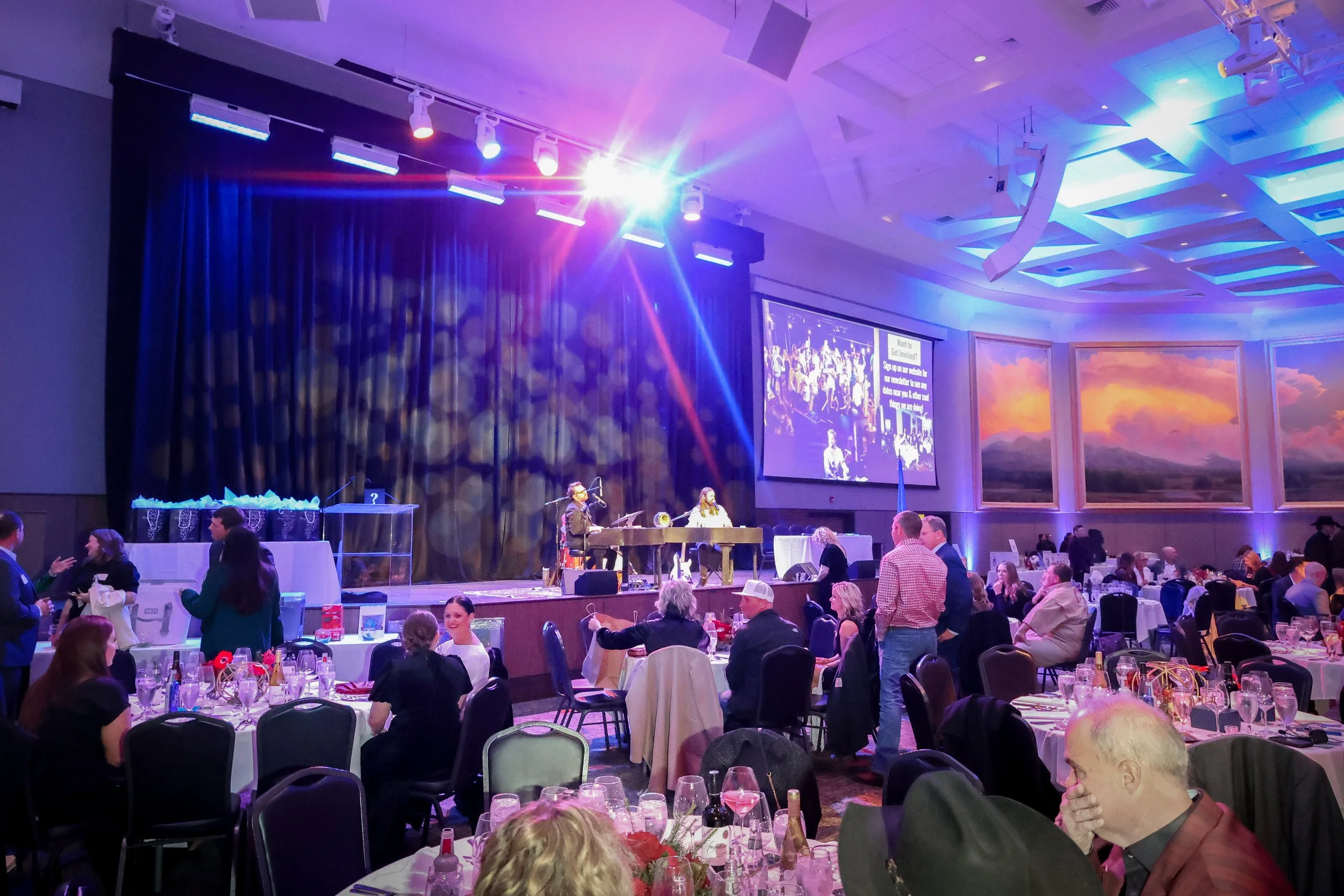 A large banquet hall with round tables set for an event, featuring floral centerpieces, glasses, and tableware. There is a stage with musicians performing, and a large screen displaying photos and text. Guests are mingling and seated, with a scenic m
