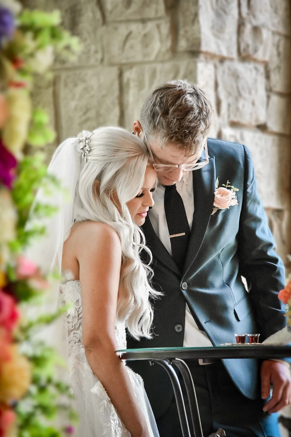 A bride and groom sharing a moment during their wedding ceremony, standing close with heads bowed, surrounded by colorful flowers and stone wall background.