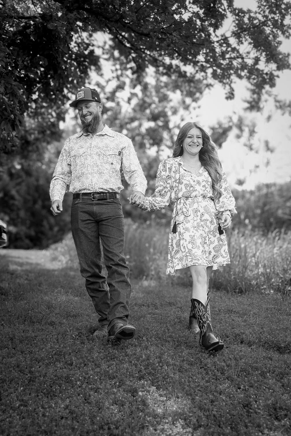 A man and woman walking hand in hand outdoors, smiling, dressed in casual clothing, with trees and grass in the background, black and white photo.