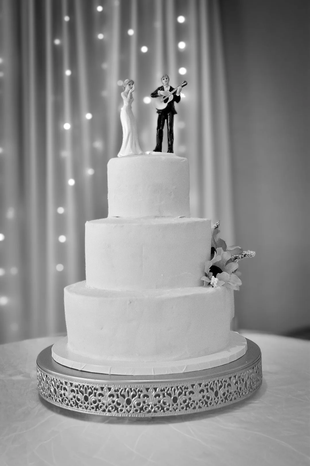 Black and white photo of a three-tiered wedding cake with floral decorations and a topper of a bride and a groom playing guitar, set against a backdrop of draped curtains and lights.