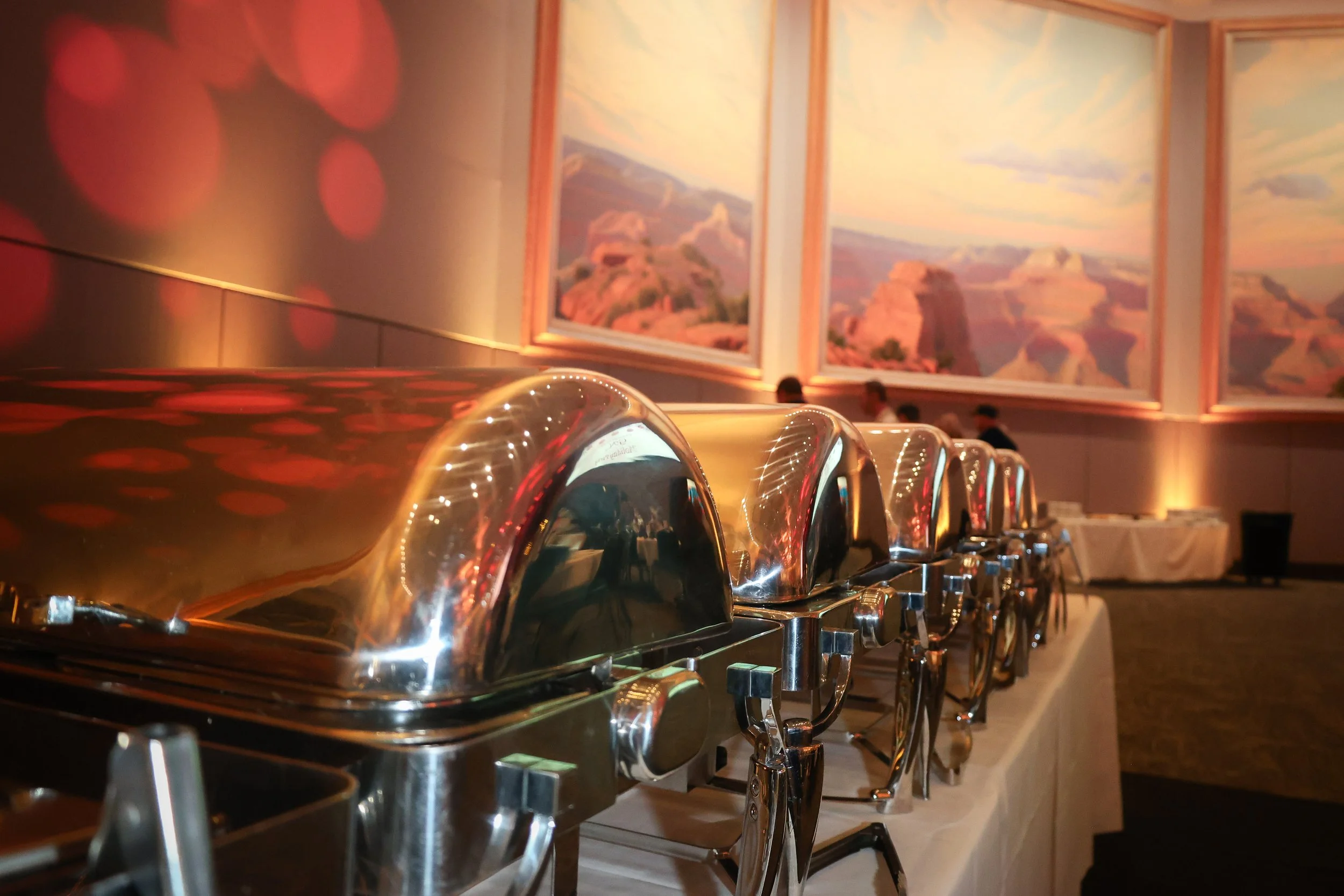 Buffet chafing dishes on a table in a banquet hall with large landscape paintings of the Grand Canyon on the walls.