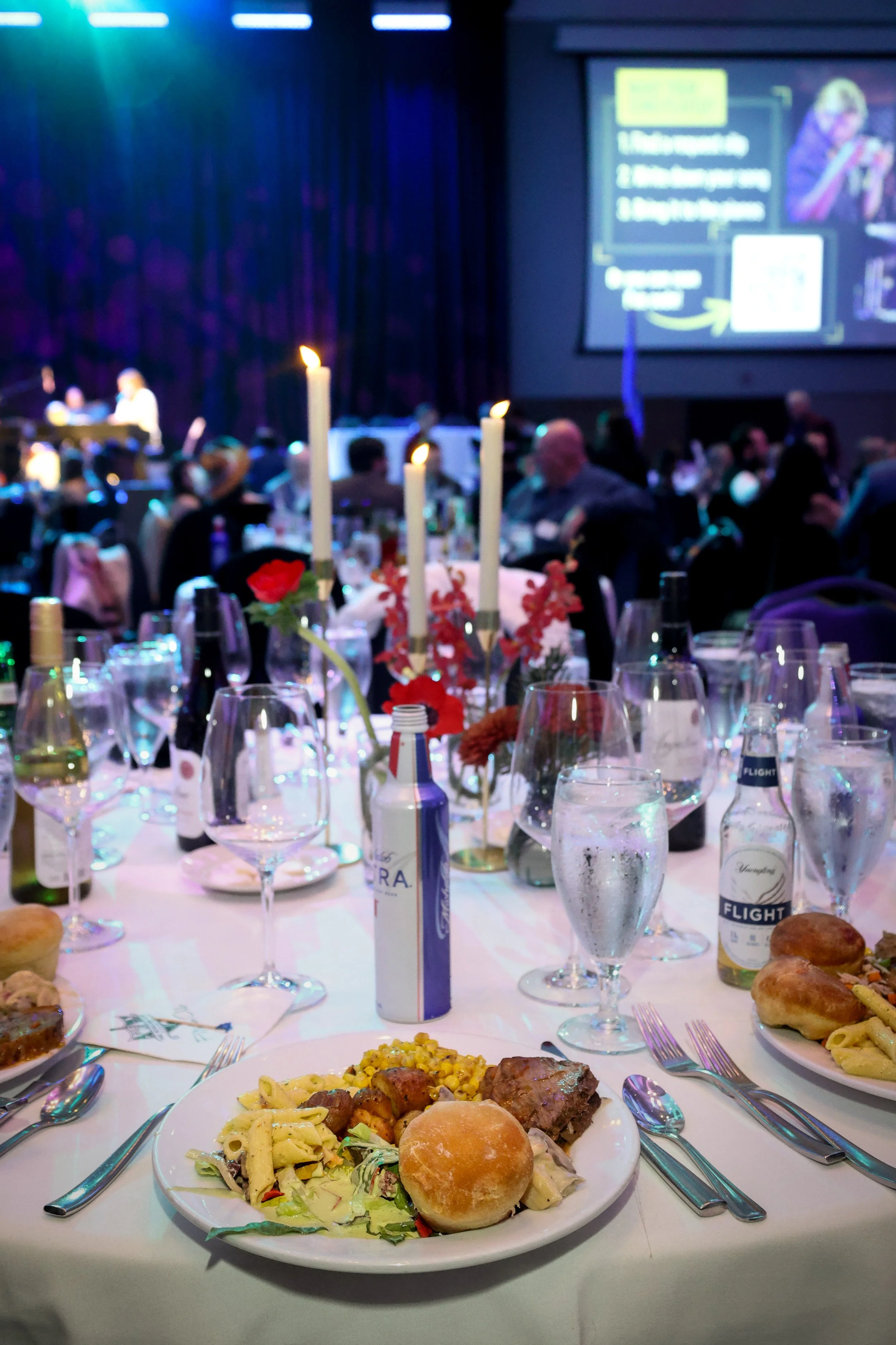 A banquet table set with plates of food, wine glasses, bottles, and a centerpiece with candles, at a formal event or celebration.