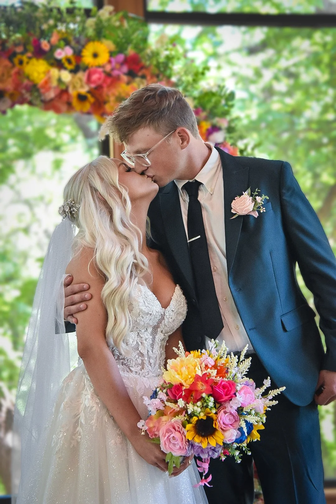 A bride and groom share a kiss during their wedding ceremony. The bride has long, wavy blonde hair with a decorative hairpiece and wears a white lace wedding gown. The groom wears glasses, a dark suit with a light shirt, black tie, and a boutonnière. The bride holds a colorful bouquet of flowers, and there are vibrant flowers in the background.