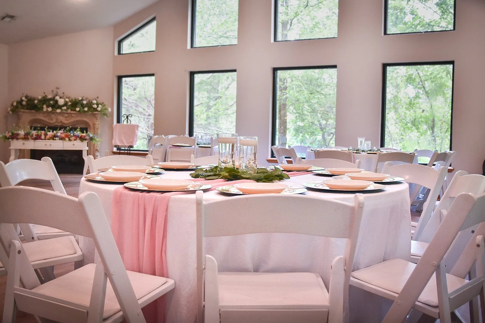 Indoor event space decorated for a celebration with round tables, white chairs, pink table runners, and views of green trees through large windows.