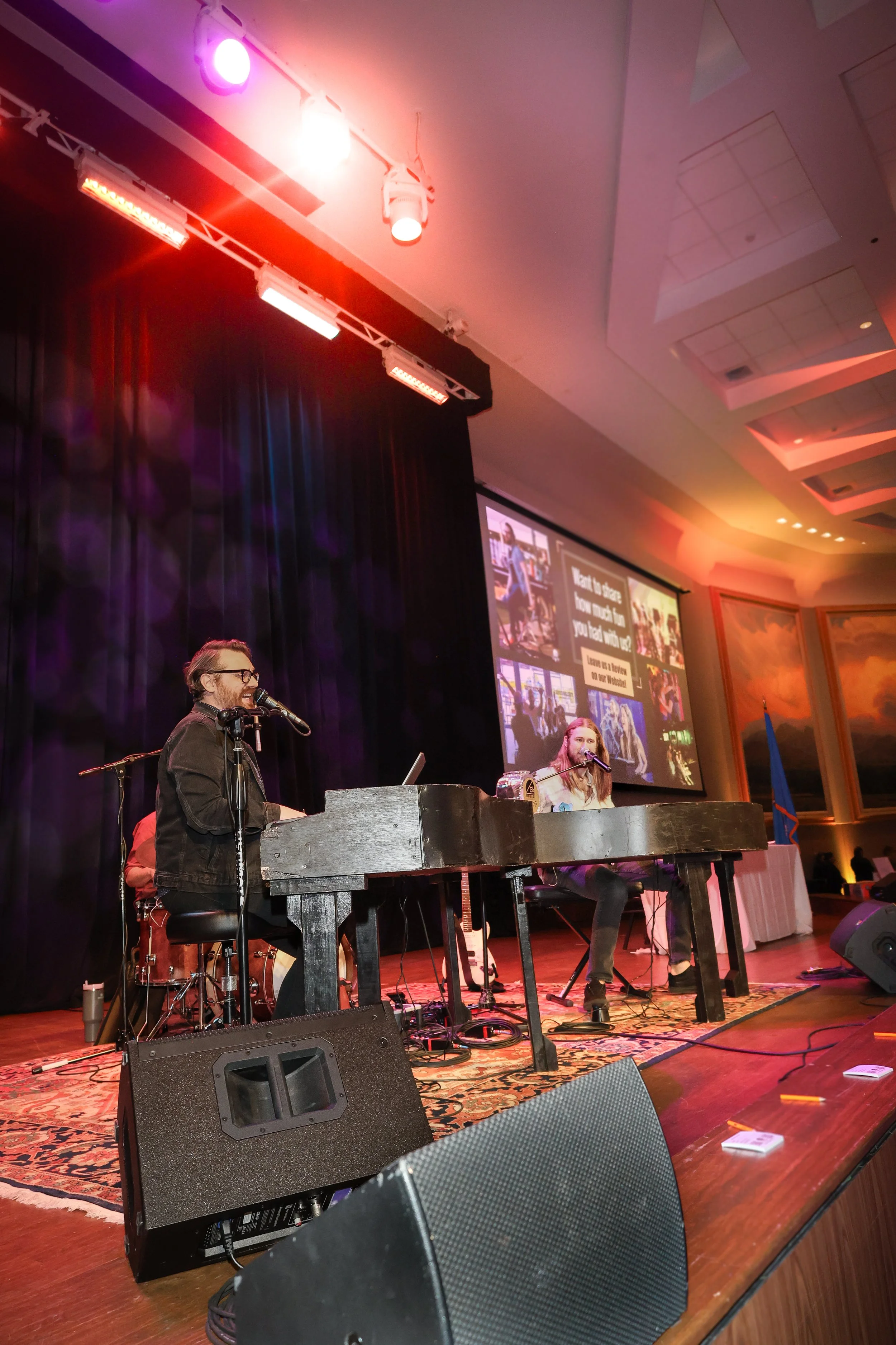 Two musicians performing on stage with a keyboard and microphone, with a drum set in the background, under stage lights, and a large screen displaying a presentation behind them.