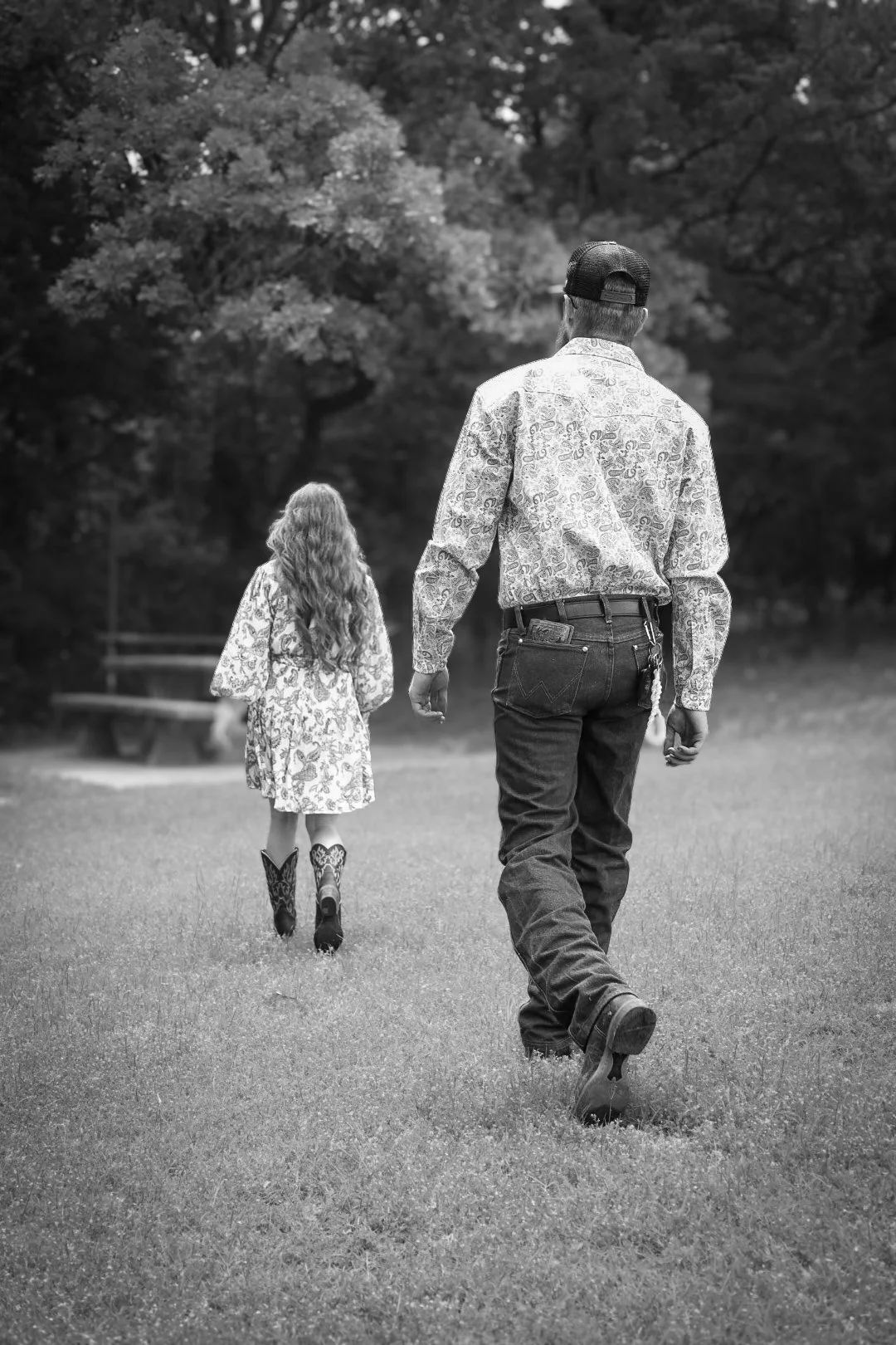 A man and a girl walking away in a park, with the man on the right and the girl on the left, surrounded by trees and grass, in black and white.