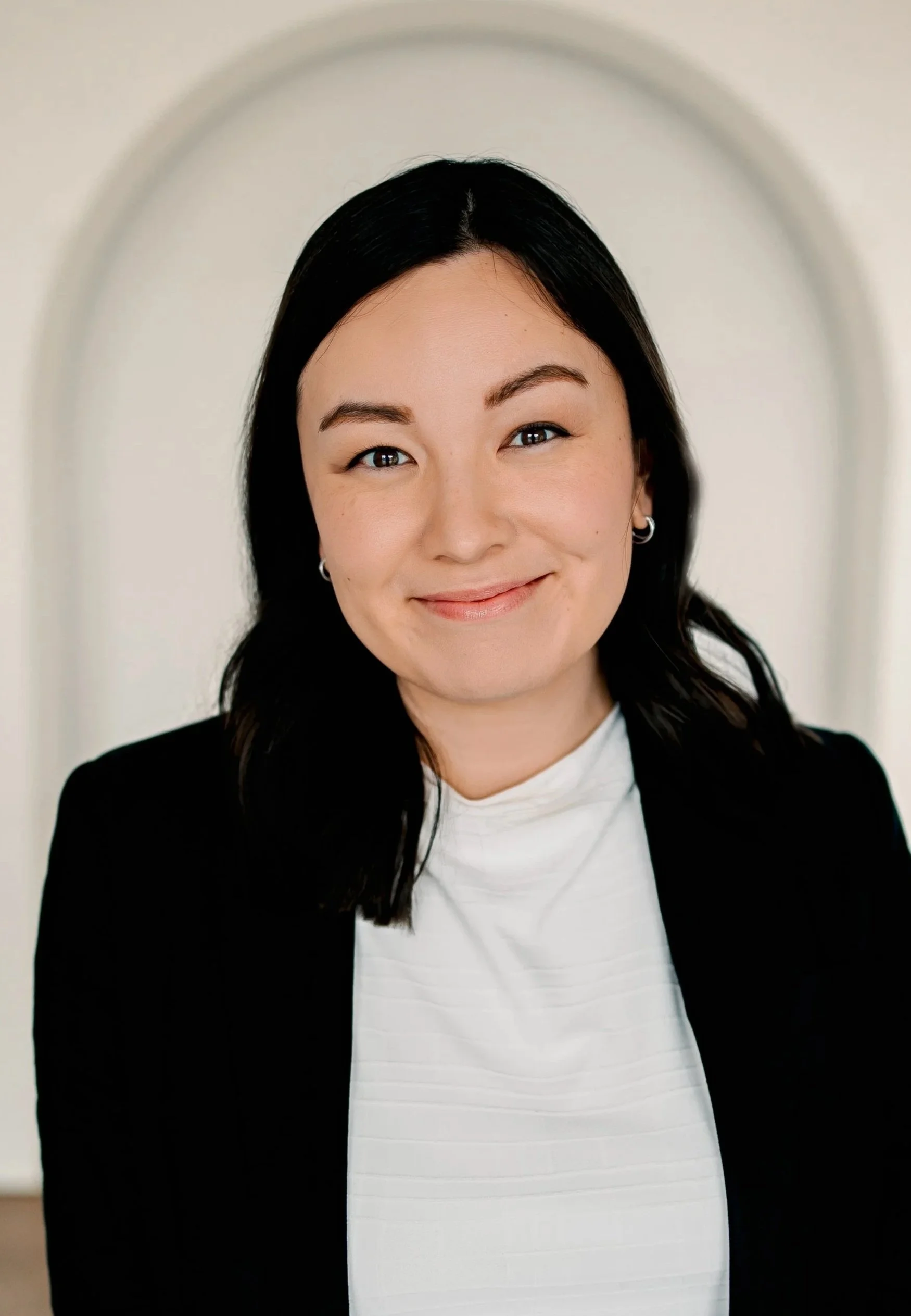 A woman with black hair, wearing a black blazer over a white top, smiling at the camera against a neutral background.