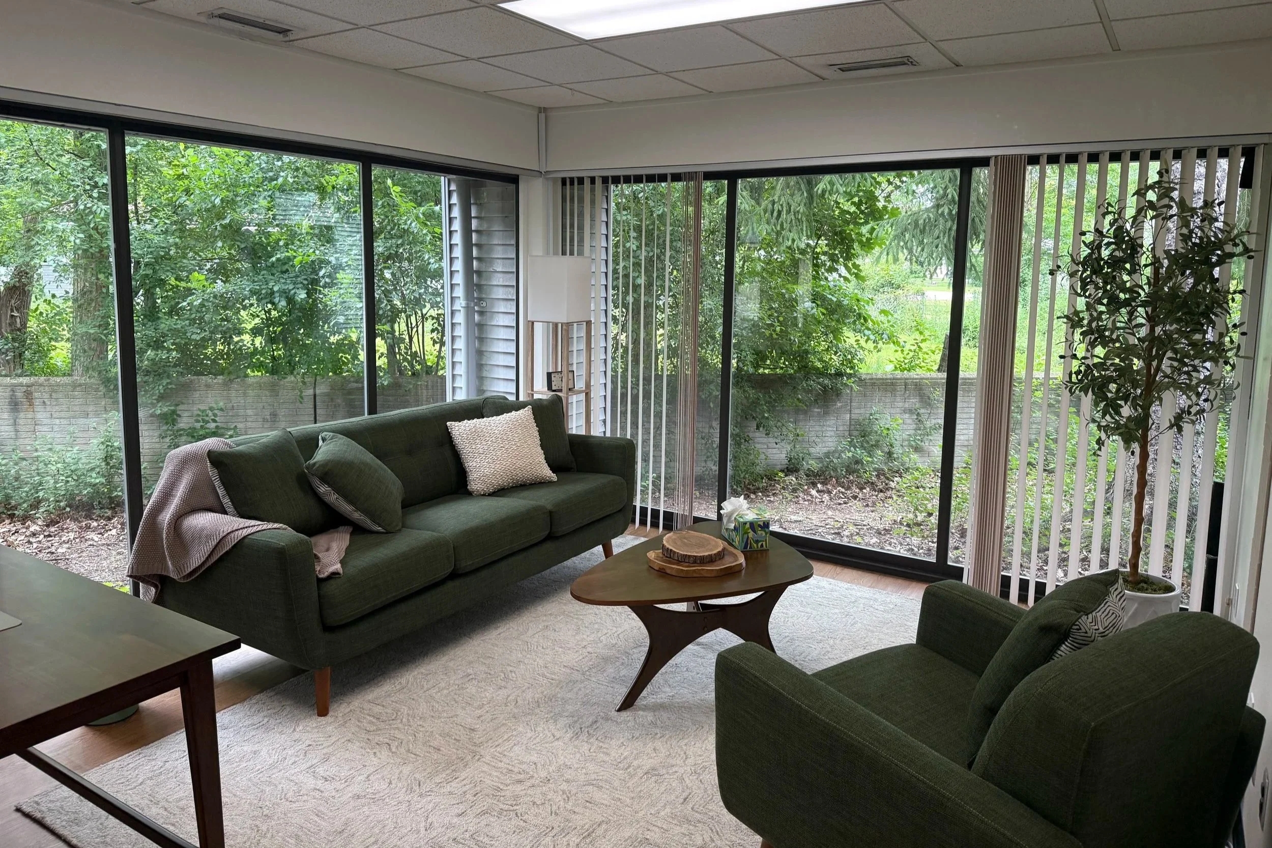 A living room with large floor-to-ceiling windows revealing green trees outside. There are green armchairs, a sofa with pillows, a wooden coffee table with decorative items, and a small side table in the space.