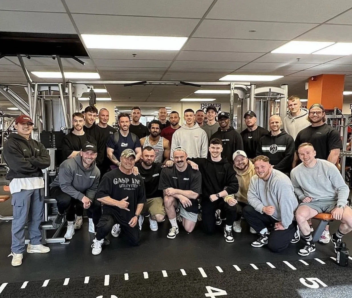 Group of people in a gym posing for a photo, some in workout clothes, with gym equipment in the background.
