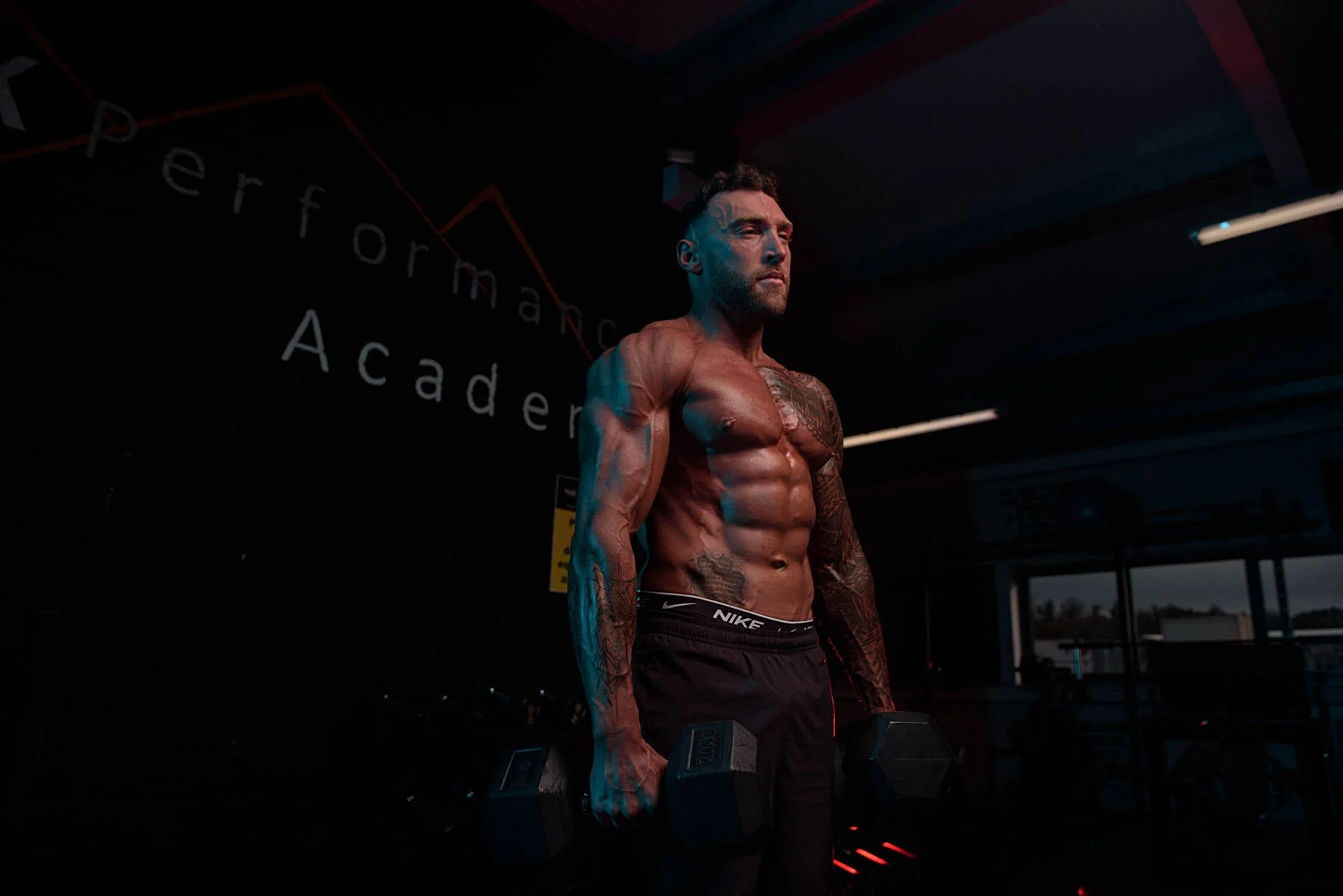 A muscular man with tattoos lifting dumbbells in a gym with a sign reading 'Performance Academy' in the background.