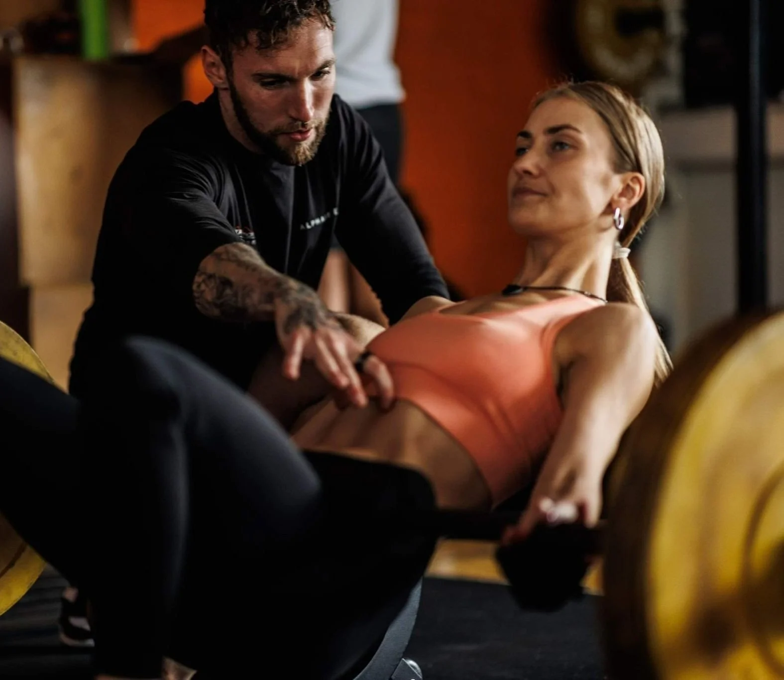 A woman in athletic wear is lifting a barbell while a trainer assists her in a gym setting.