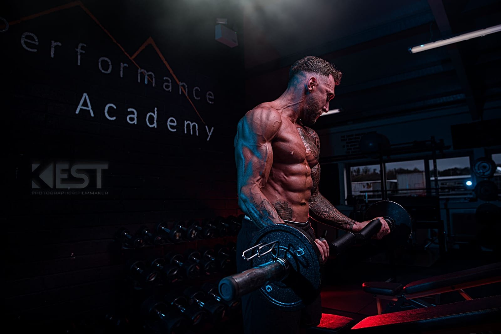 A shirtless, muscular man with tattoos lifts a barbell in a gym with dark lighting, with the words 'performance Academy' on the background wall.
