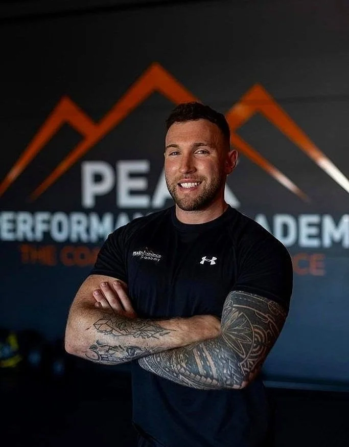 A smiling man with tattoos on his arms standing with arms crossed in front of a black background with orange mountains and white text that reads 'PEAK PERFORMANCE ACADEMY'.