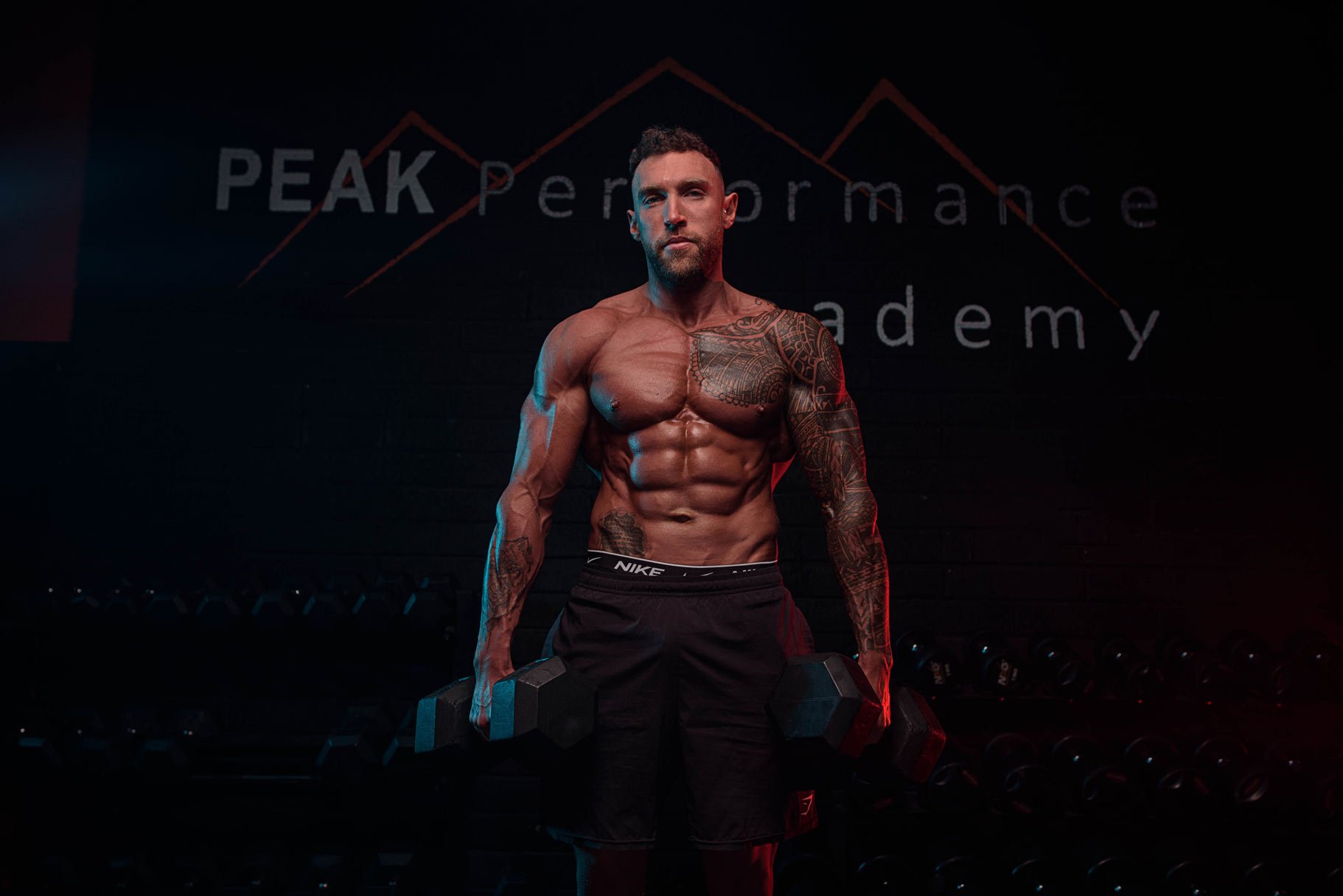 A muscular man holding dumbbells in a gym with a sign that reads 'PEAK Performance Academy' in the background.