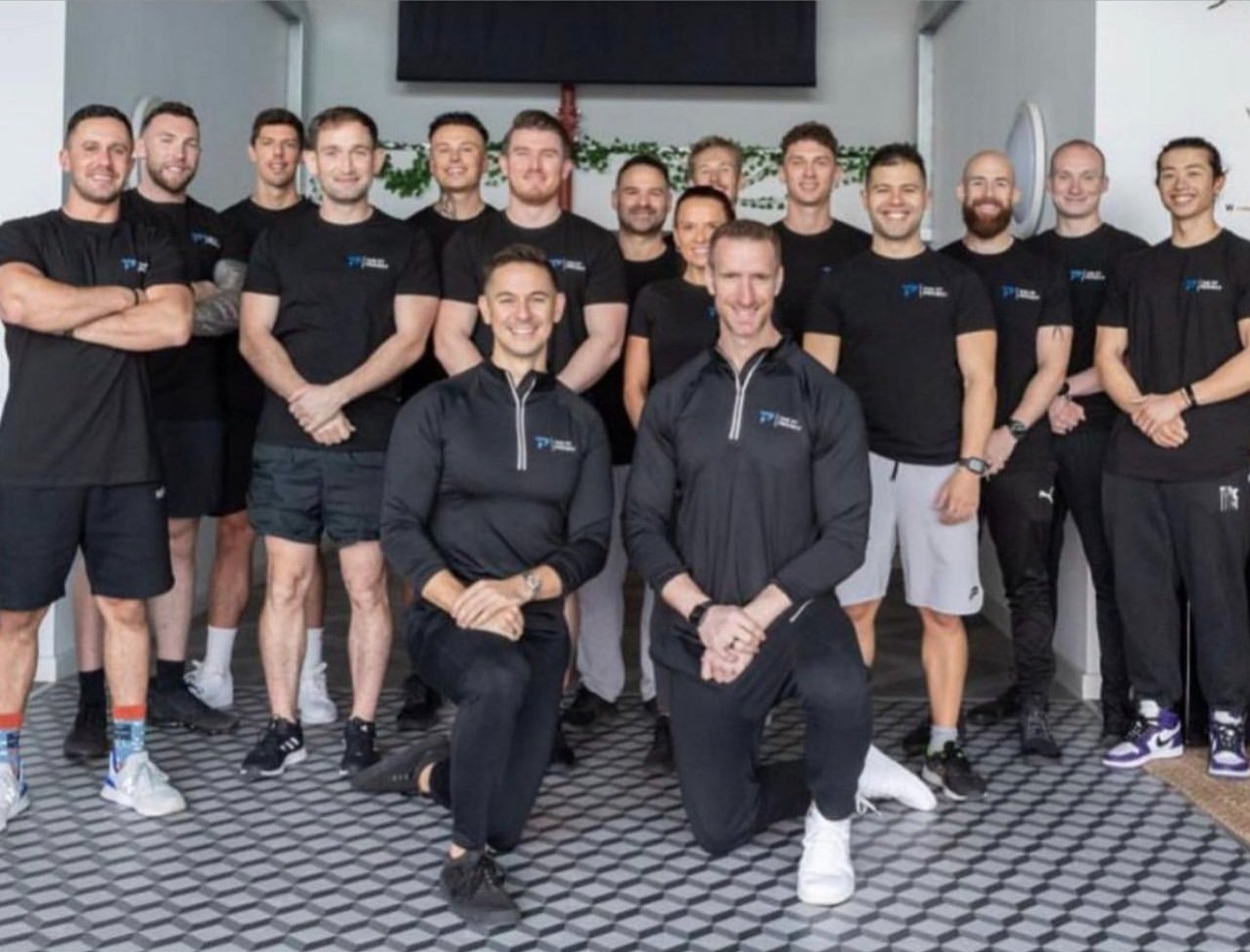 Group of 16 people posing indoors, most wearing black t-shirts and athletic wear, some kneeling in front, others standing behind, smiling, with a striped carpet floor and a decorated wall behind them.