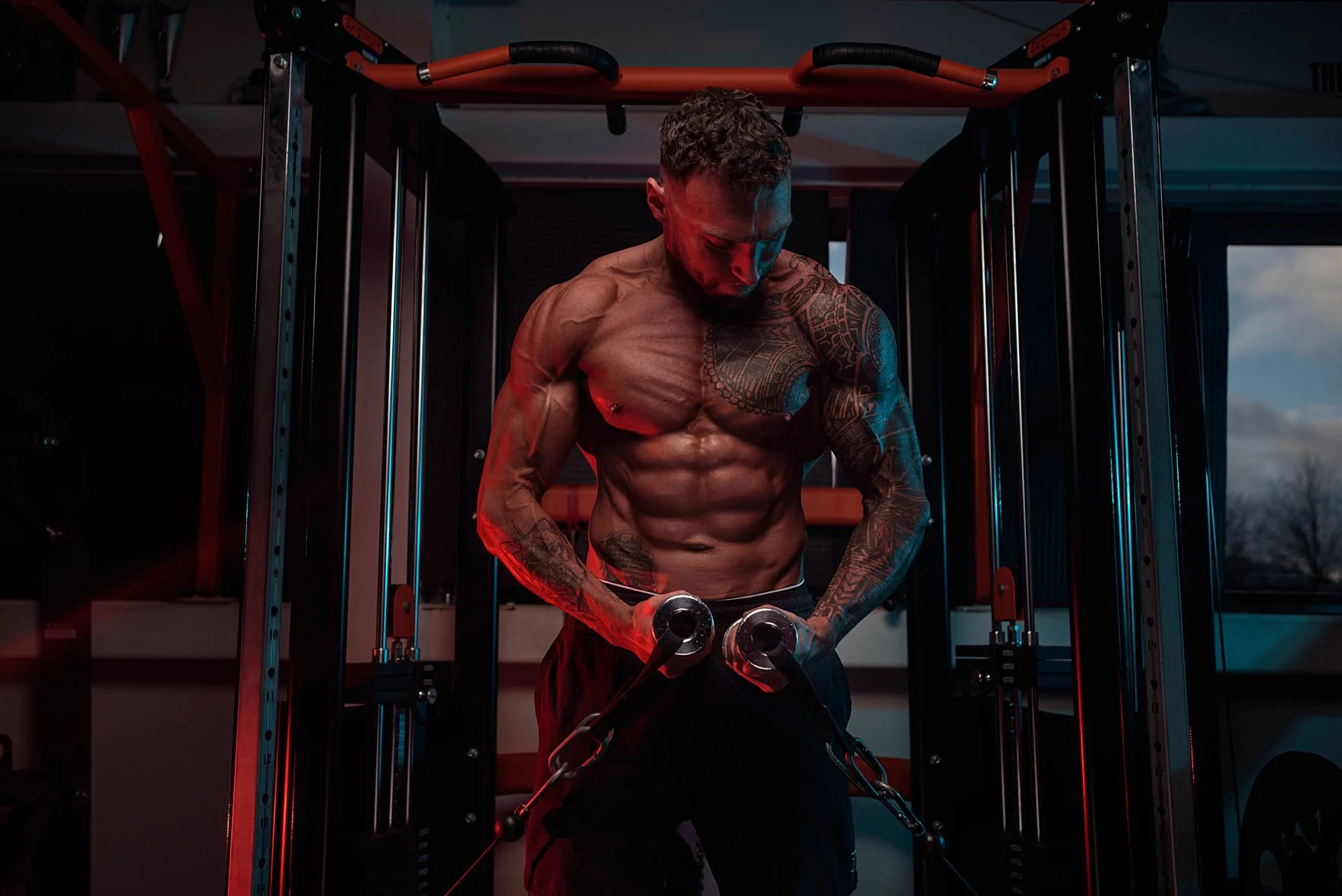 A muscular man with tattoos on his arms and chest working out with resistance bands in a gym during dusk or night.