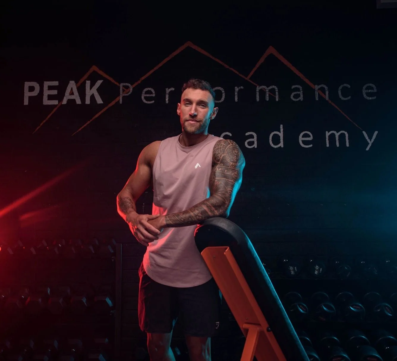 A muscular man with tattoos on his arms in a gym, standing near a workout bench, with the sign 'PEAK Performance Academy' in the background.