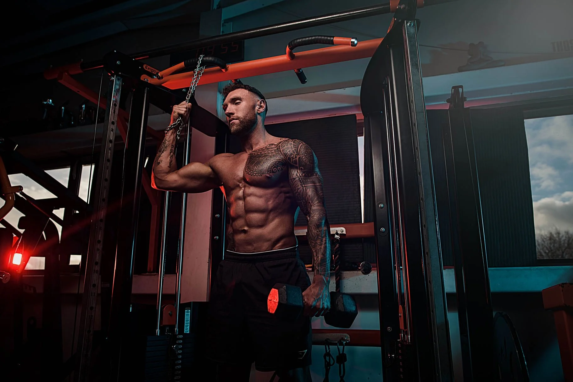 A muscular man with tattoos on his right arm lifting a dumbbell in a gym, holding a chain with his left hand, with gym equipment and windows showing a cloudy sky in the background.