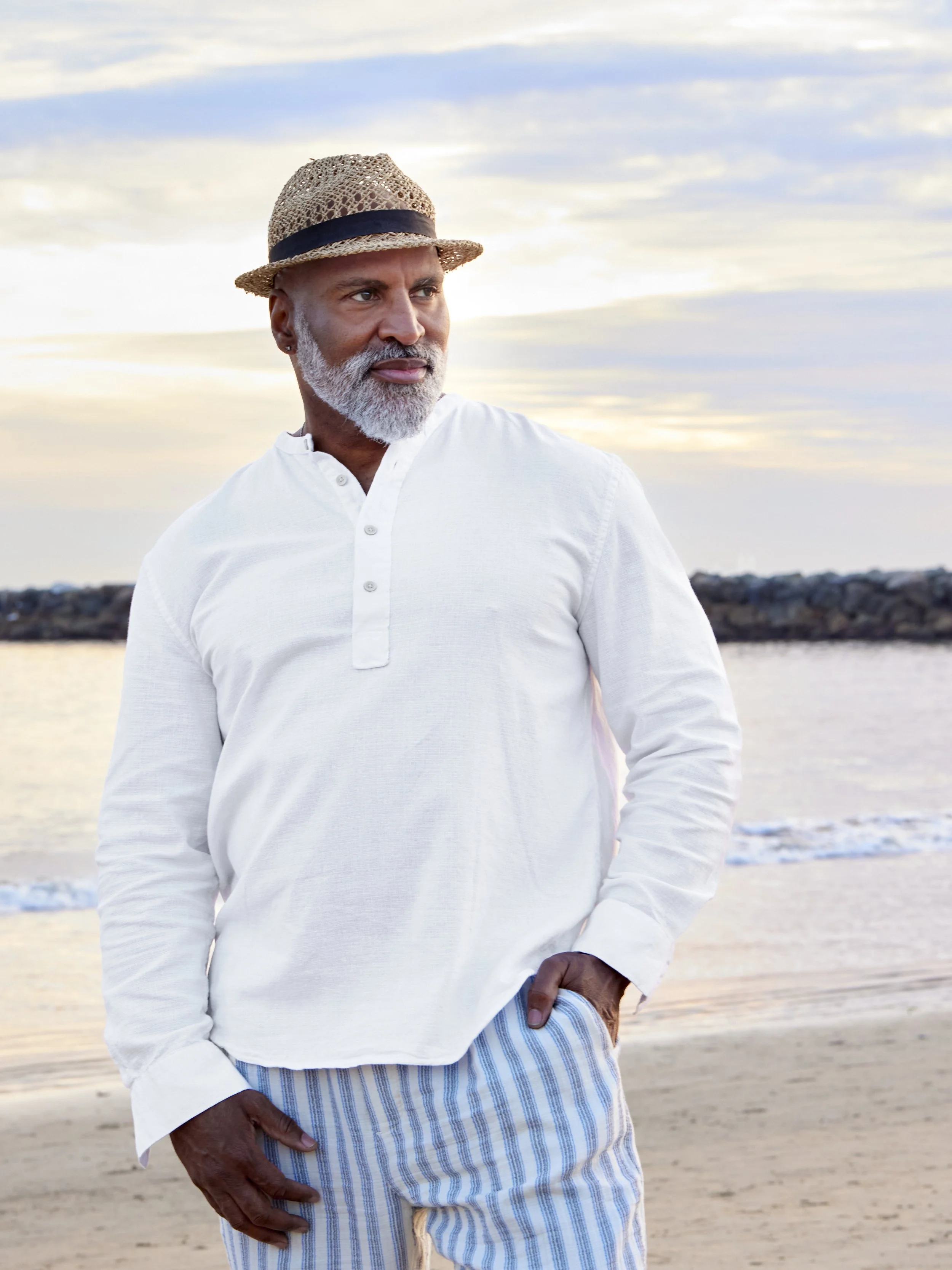 Elegant mid‑life sophistication meets sun‑kissed resort charm: a distinguished gentleman clad in a crisp ivory linen Henley, paired with soft blue‑and‑white striped linen trousers and a natural straw fedora, stands poised at the water’s edge under a 