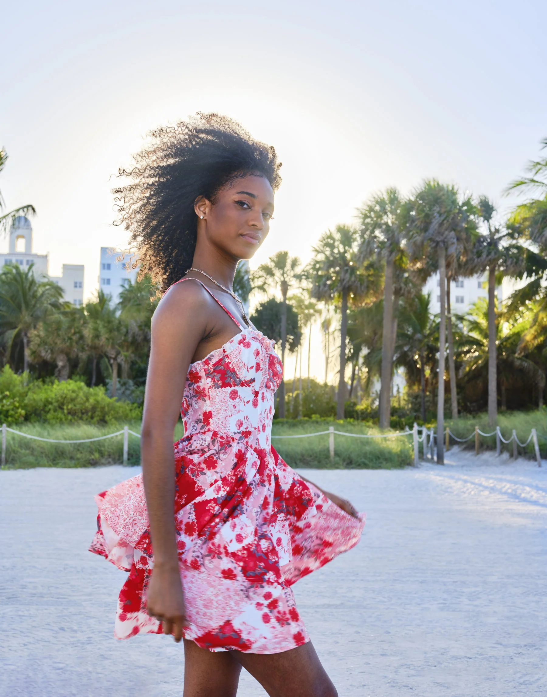 A striking destination‑resort moment: a confident young woman with natural curls turns gracefully in a flowing red and white floral summer dress, the golden light of sunset haloing her silhouette as palm trees and coastal architecture form a chic bac