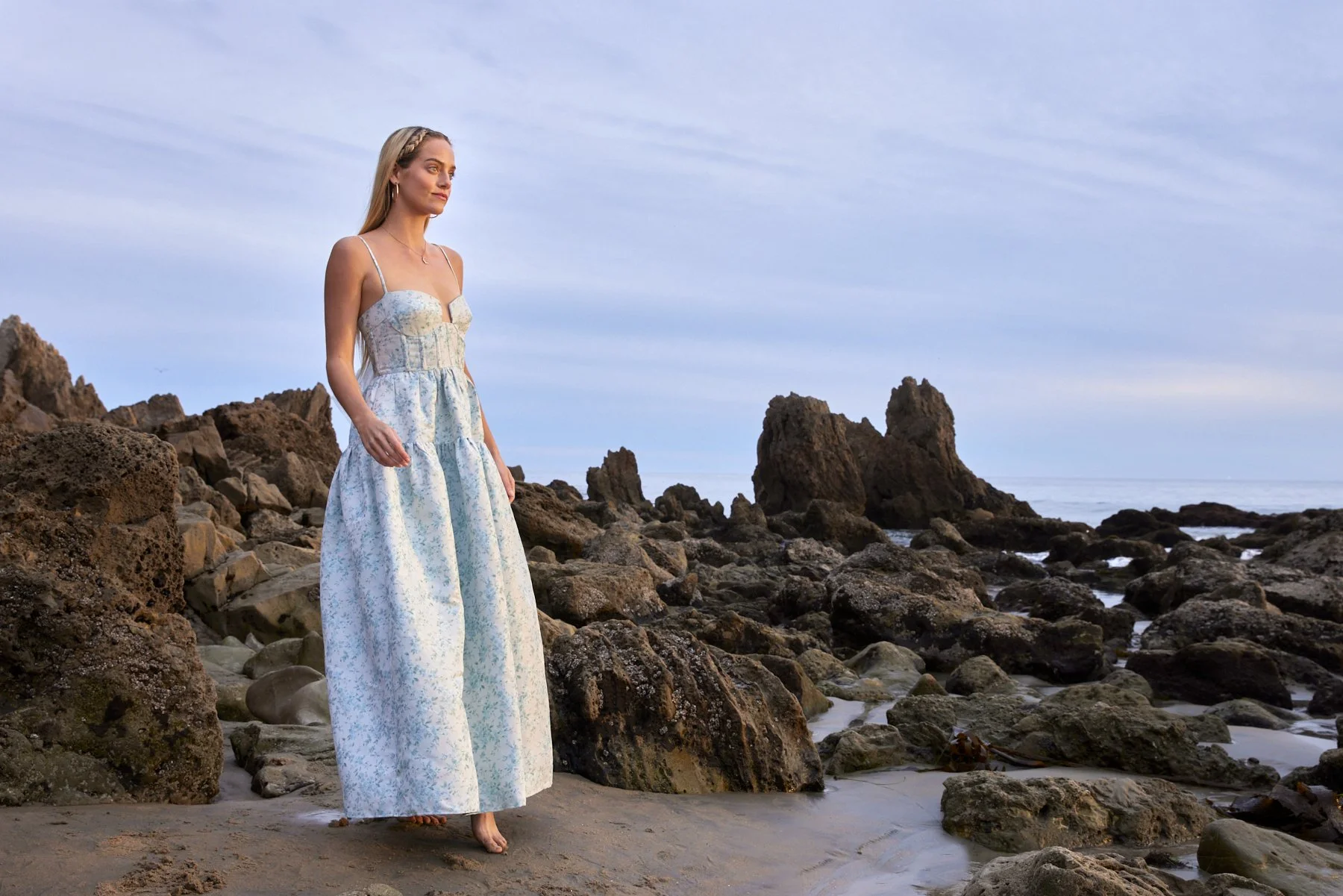 A serene moment of modern elegance: A luminous, barefoot model in a soft‑blue, floral‑patterned couture gown walks across rugged coastal rocks at dusk, embodying refined style and effortless grace. Set against the dramatic seaside landscape of a luxu