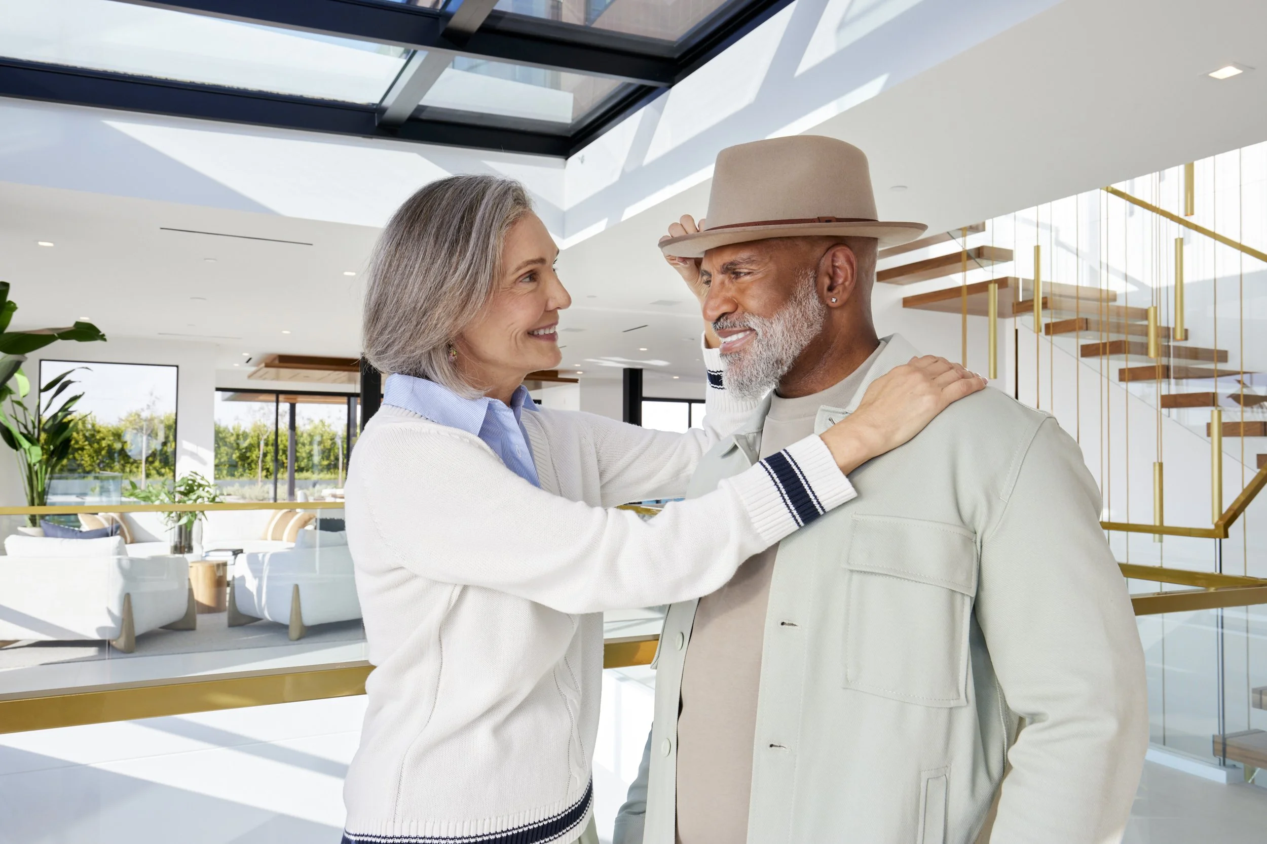 In this elegant editorial moment, a stylish couple shares a warm smile in a serene, sun‑filled modern loft, perfectly blending timeless sophistication and relaxed luxury. She wears a classic ivory cashmere sweater layered over a crisp light‑blue Oxfo