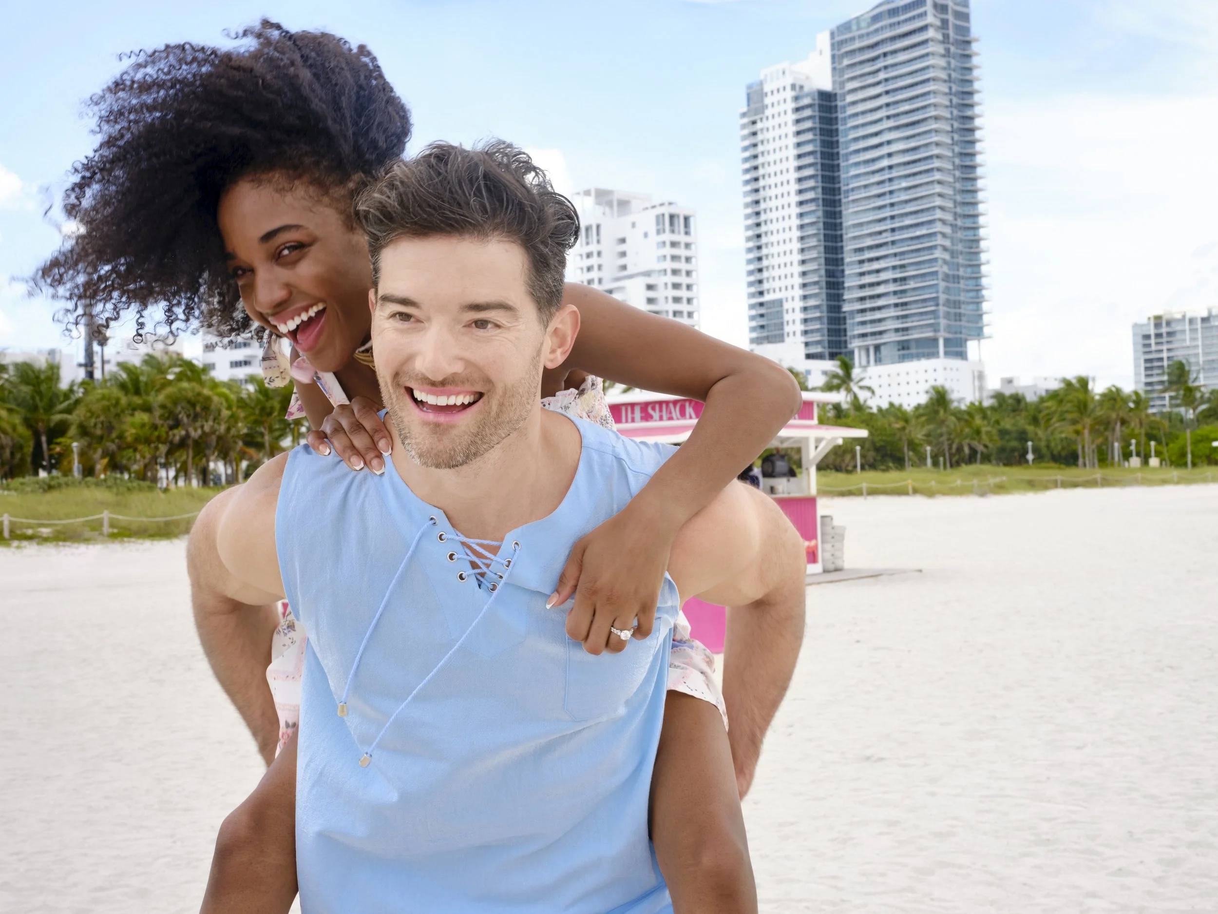 Playfully elevated at the edge of paradise — a radiant couple relishing a sun-lit moment on the white-sand beach of a chic coastal resort, with swaying palms, turquoise surf, and sleek urban towers rising behind them. She perches on his back, her wil