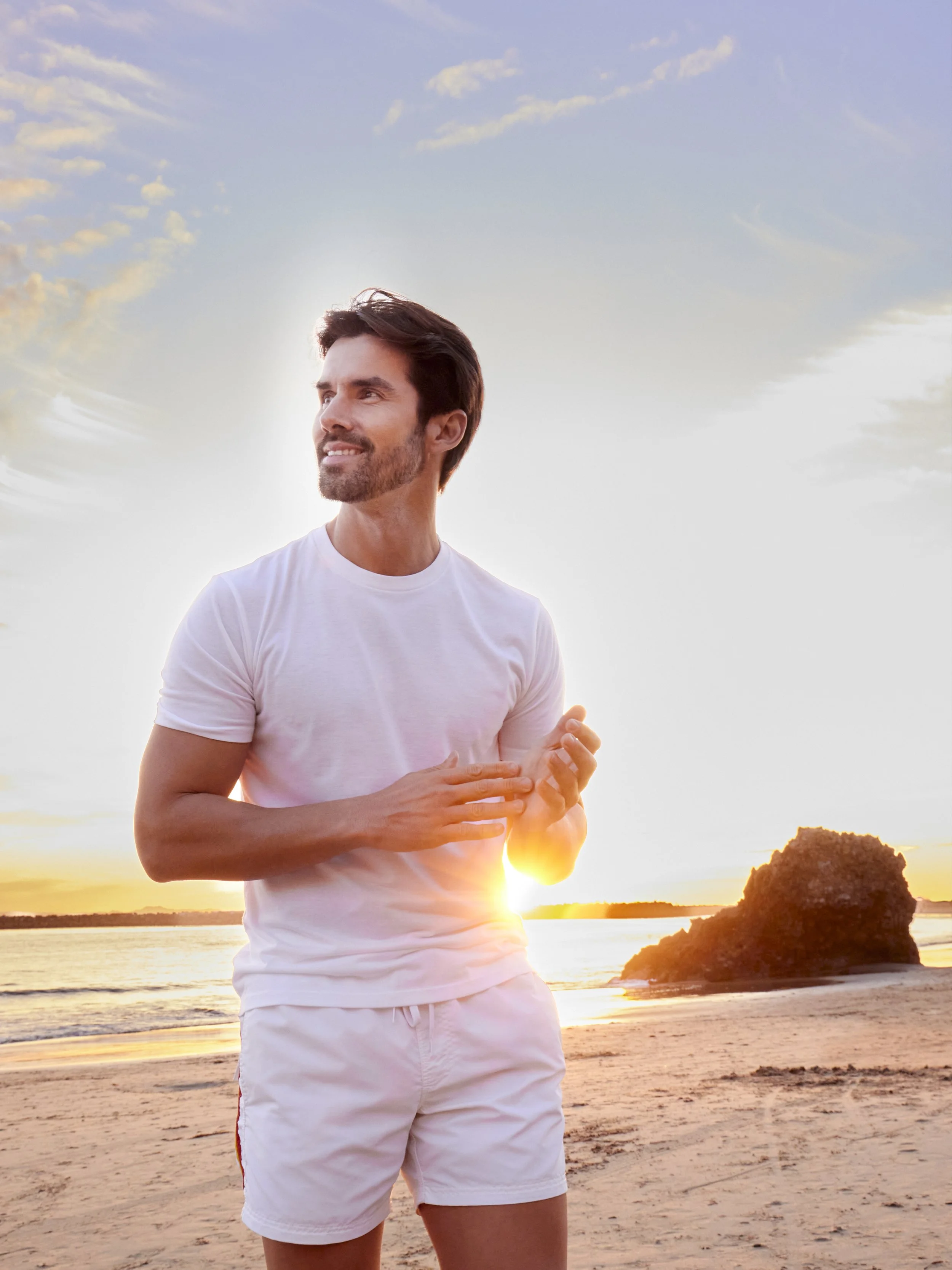 A refined lifestyle portrait of a modern gentleman posing at sunset on a pristine beachfront at a luxury resort destination. Dressed in crisp white shorts and a tailored white tee, he embodies effortless high‑end resort style and fashion editorial fl