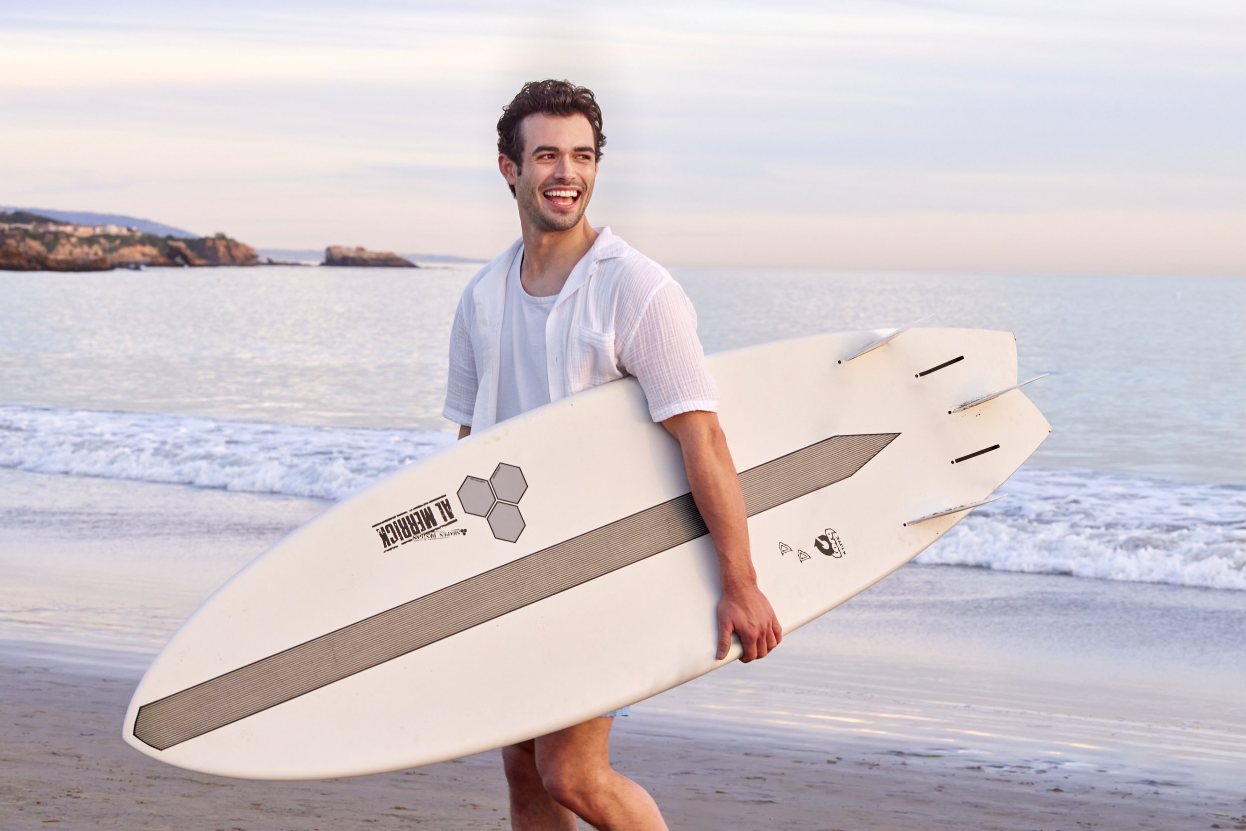 A radiant sunset glow bathes this seaside moment: a stylish, effortlessly confident man strides along the shoreline, surfboard in hand, embodying the refined spirit of coastal luxury. With an open‑weave linen shirt layered over a crisp white tee, he 
