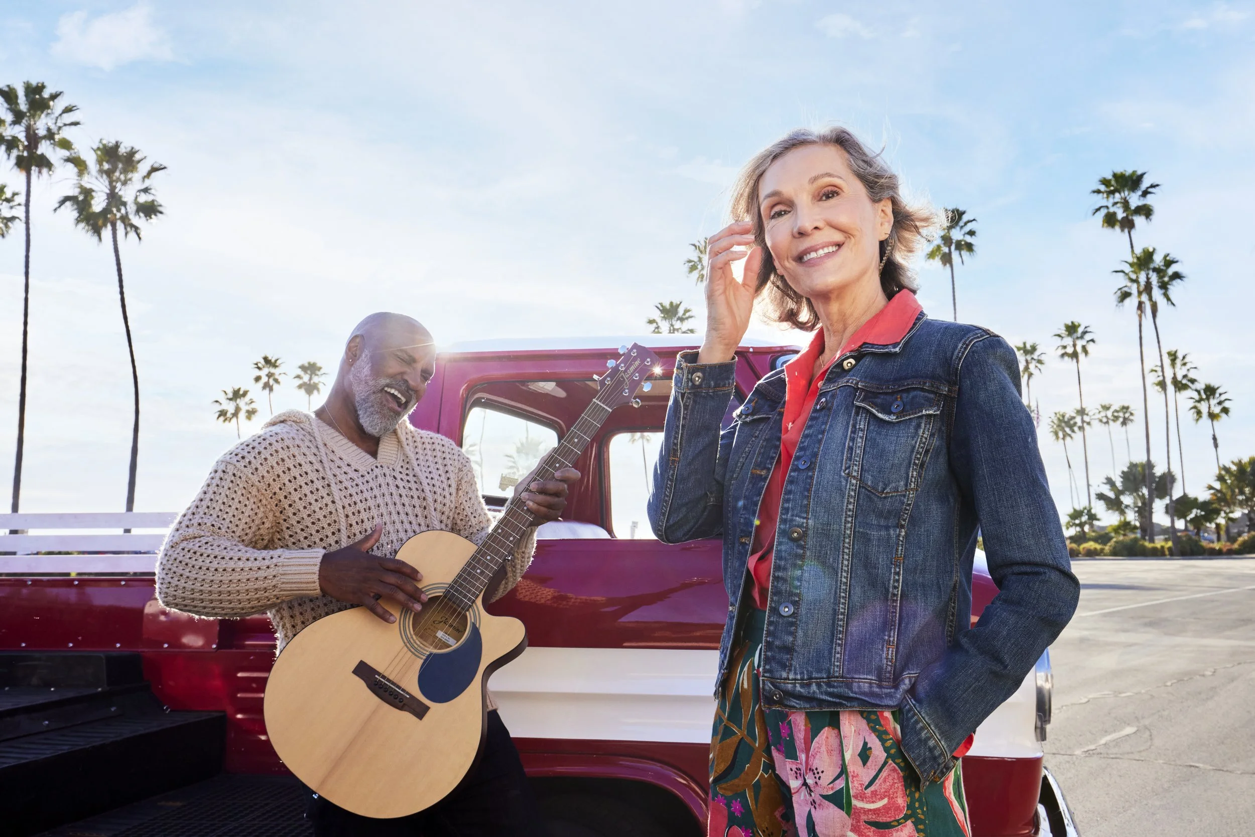 A joyful couple enjoys a relaxed outdoor moment by their vintage red-and-white pickup truck on a sunlit street lined with palm trees. The older man strums an acoustic guitar, while the stylish woman, wearing a denim jacket and a colorful patterned sk