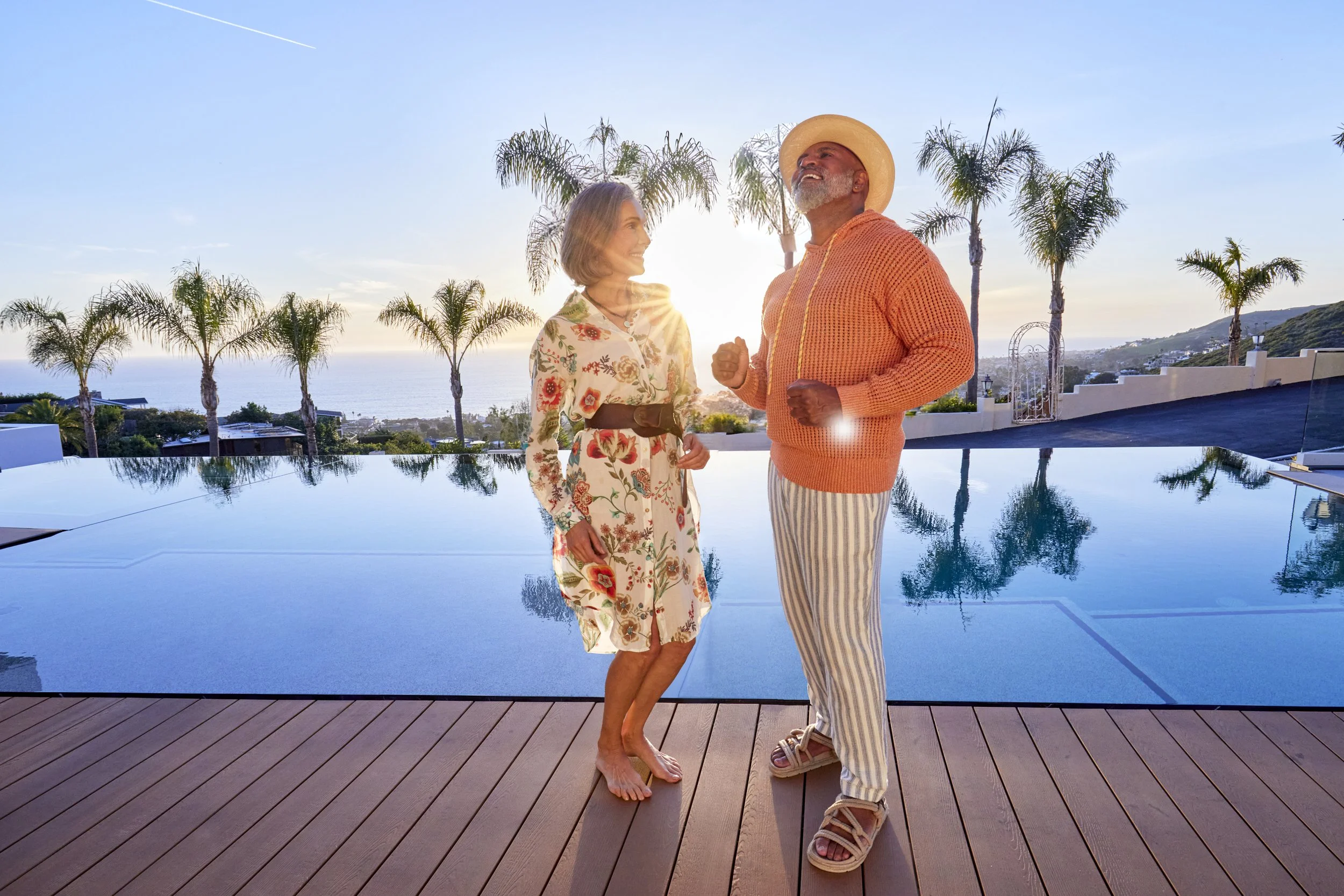 A stylish man and woman stand barefoot on a sleek teak pool deck at the edge of a modern infinity pool, as the sun sets over the Pacific horizon. The woman wears a light floral‑print shirt‑dress cinched with a wide leather belt, while the man is dres