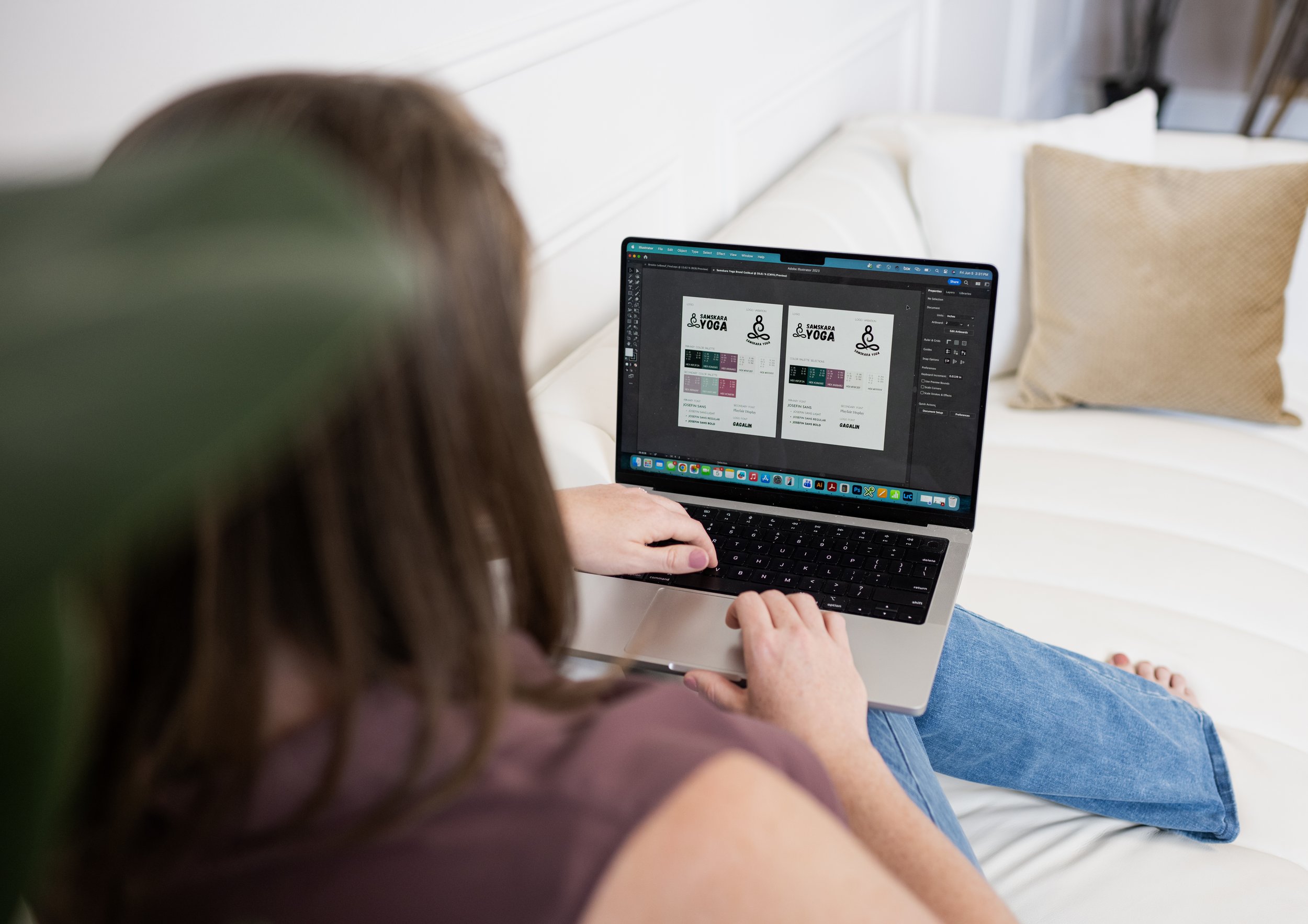 Close-up of a woman with curly hair working on a laptop, displaying a design containing the words 'SAMSKARA YOGA' and yoga-related symbols on the screen, sitting on a white couch with a beige pillow.