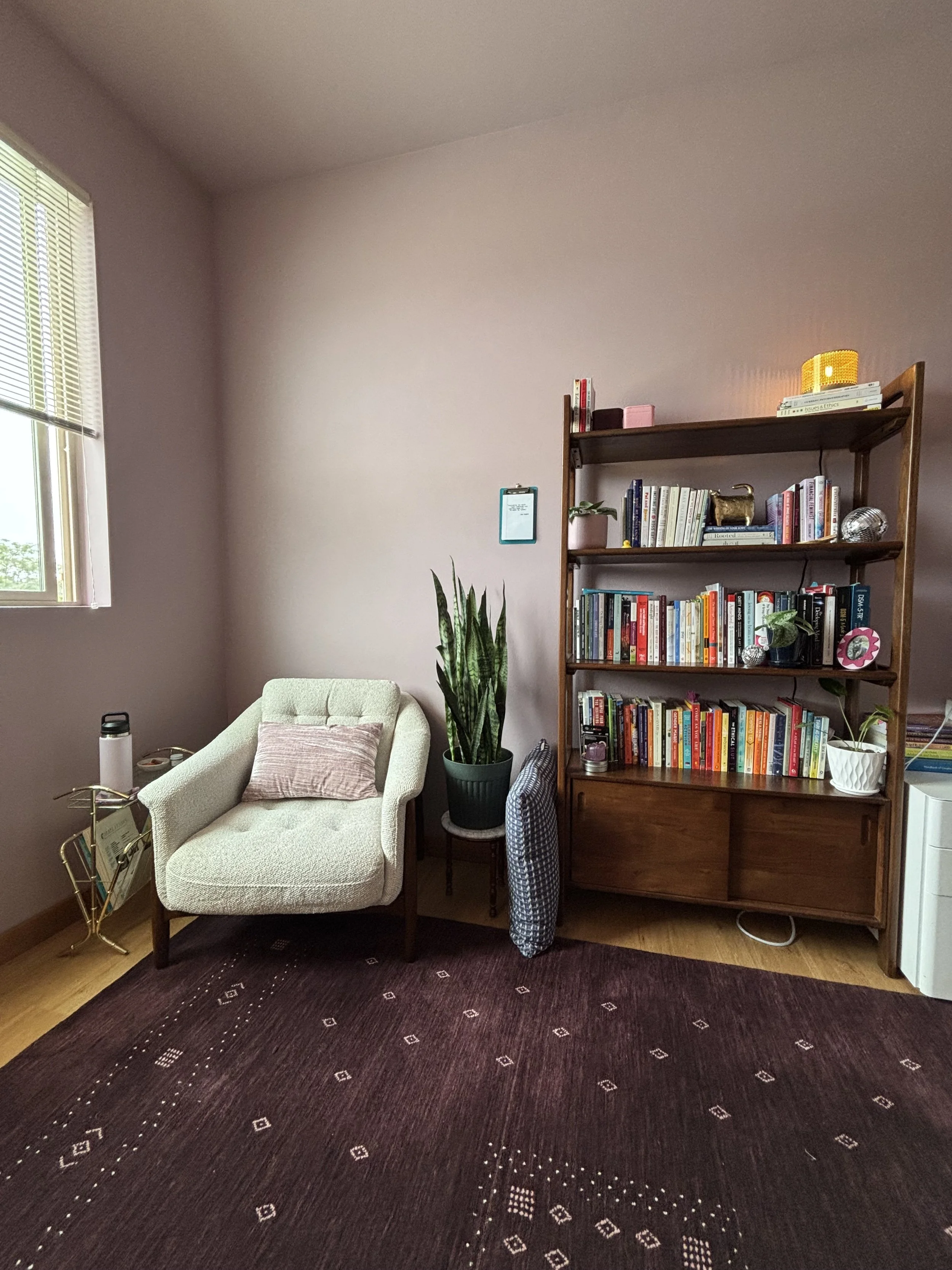 A cozy office corner with a white armchair, a pink cushion, a tall green plant in a pot, a bookshelf filled with books, and a dark patterned rug on hardwood floor next to a window with blinds.